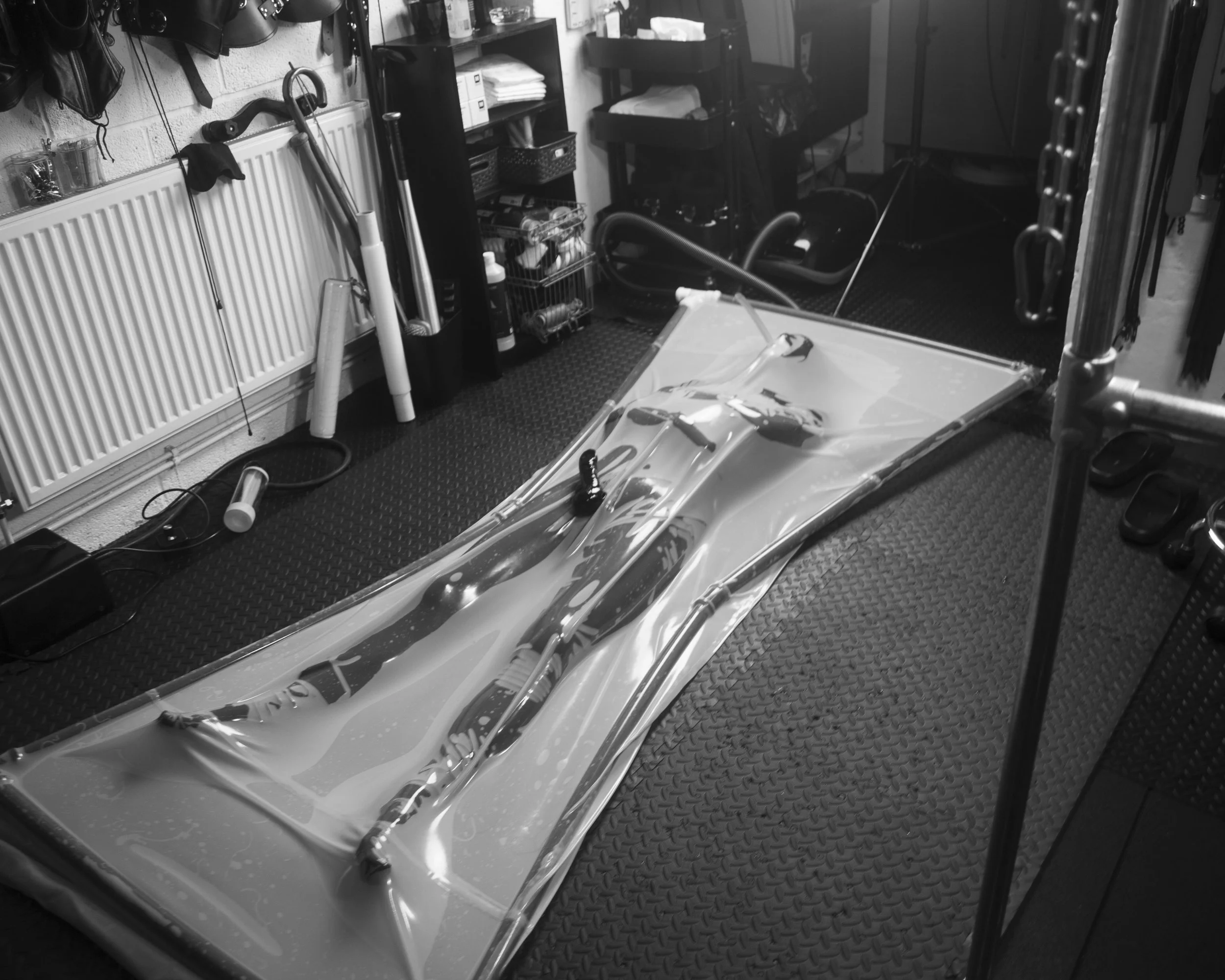 The image shows a black and white view of a garage or workshop with various tools and equipment. There are hanging helmets, a pipe wrench, and cleaning brushes on a wall-mounted rack. A small shelf holds storage baskets and some stacked items. A car 