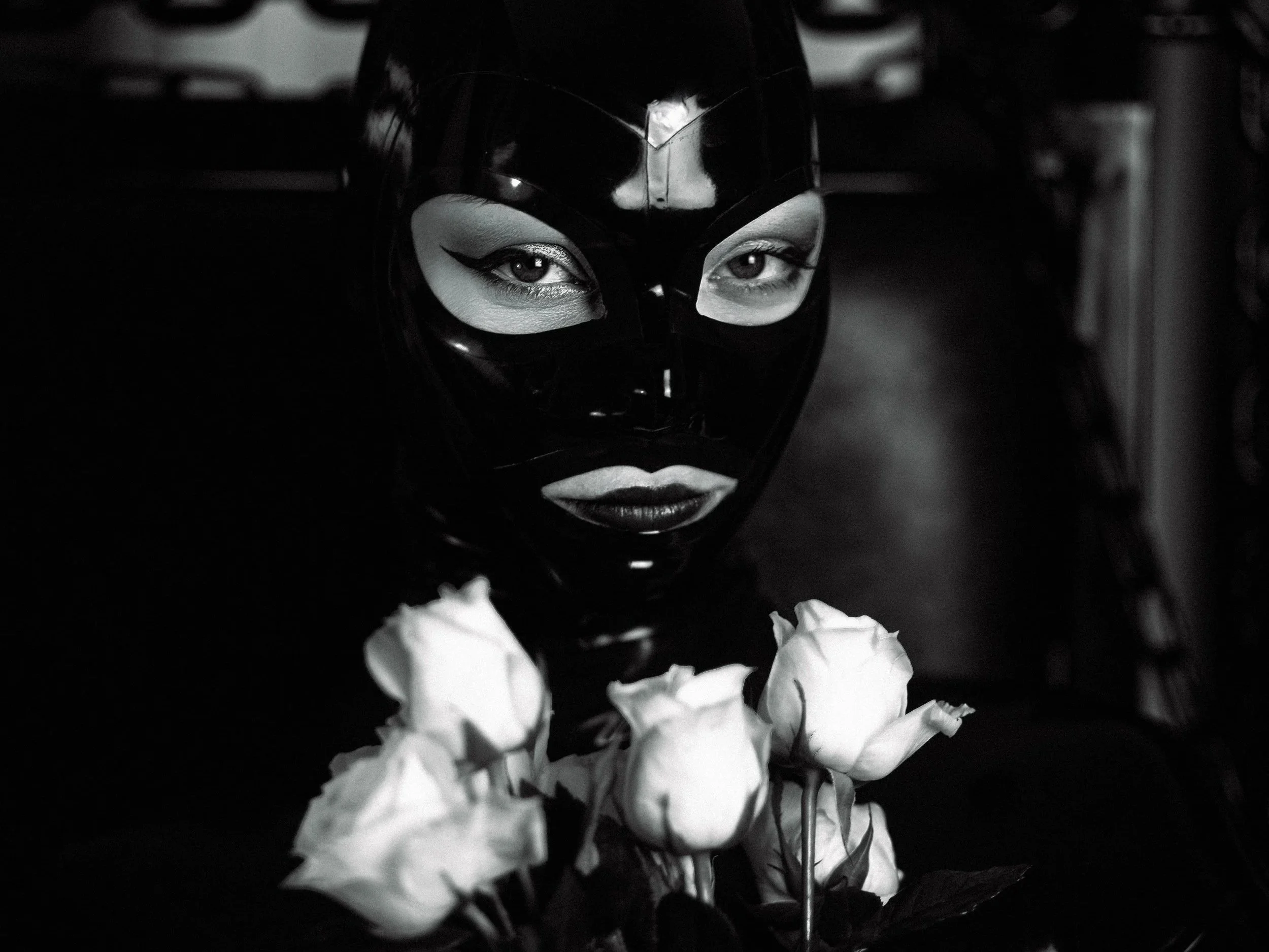 A person's face wearing a black mask with eye holes, surrounded by white flowers, in a black and white photograph.