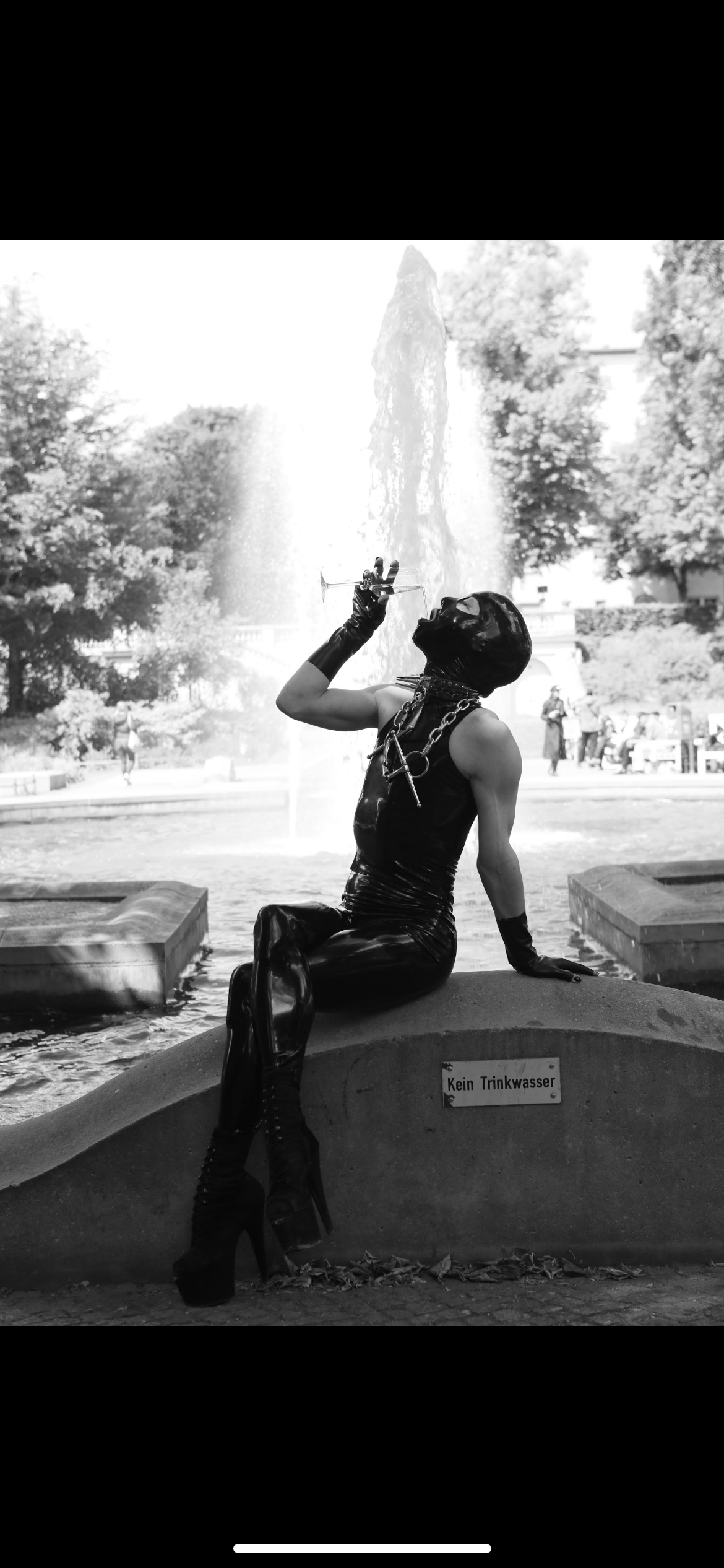 A person dressed in black, wearing a mask and high-heeled boots, sitting on a fountain with a water feature in the background, drinking from a glass. The fountain has a sign that reads "Kein Trinkwasser."