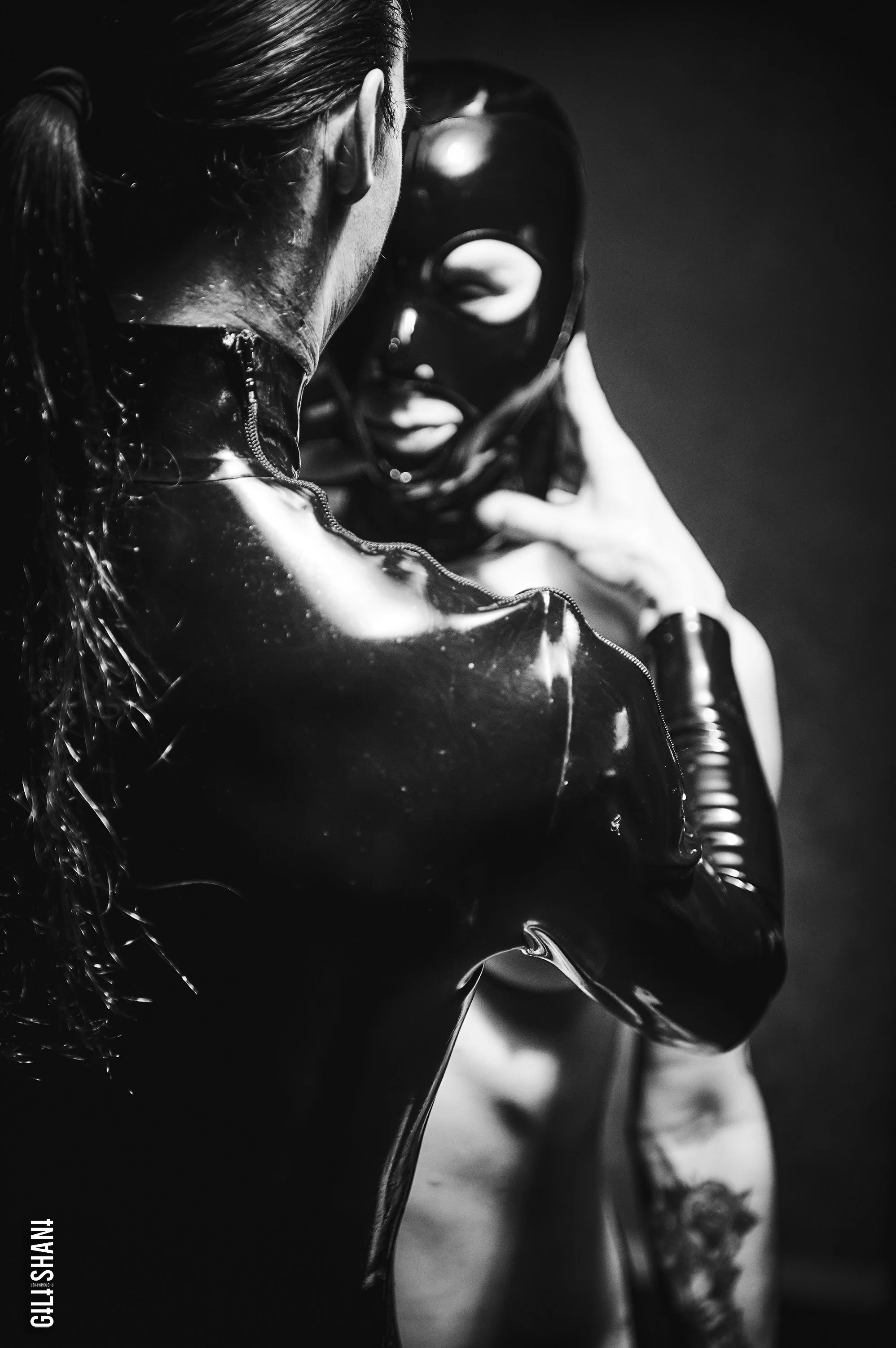 Black and white photo of two people, one wearing a shiny, black latex outfit and the other wearing a black mask with eye and mouth holes, engaging in an intimate pose.