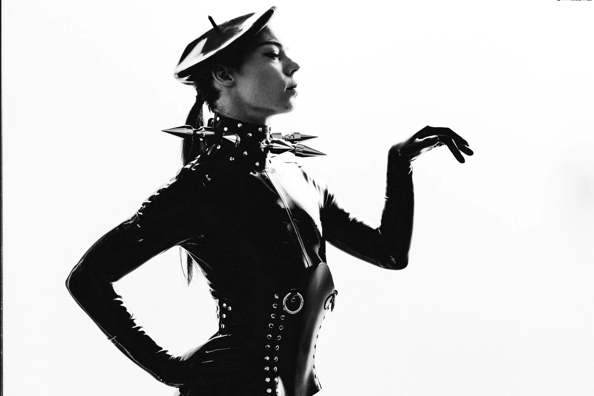 Black and white photo of a woman in a shiny, form-fitting outfit with studs, wearing a hat with a brim and a spiked choker, posing with one hand raised, in a high-contrast style.