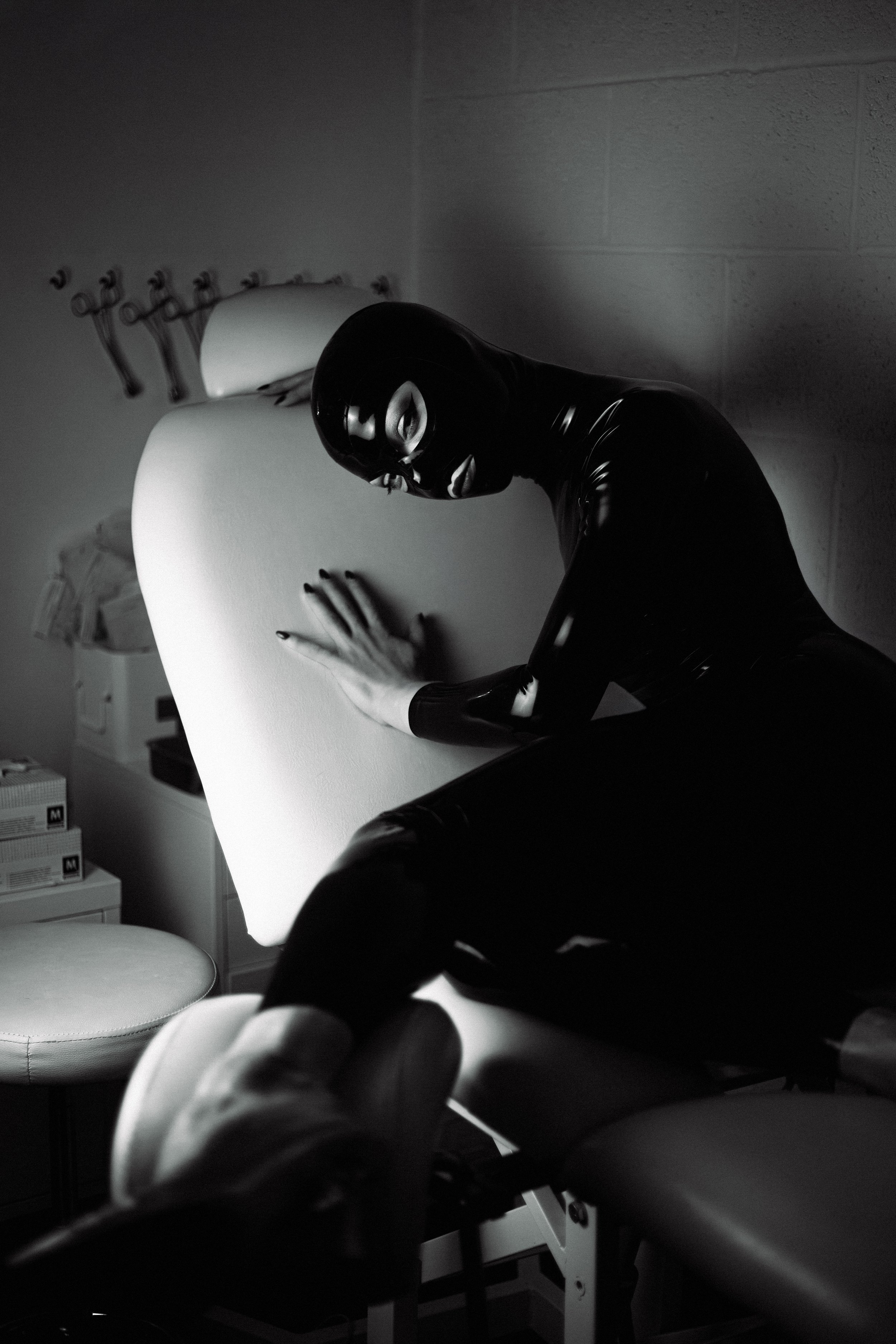 A person wearing a black bodysuit and mask, sitting in a medical or spa setting, with a white sheet or towel covering their lap and chest, surrounded by shelves and boxes.