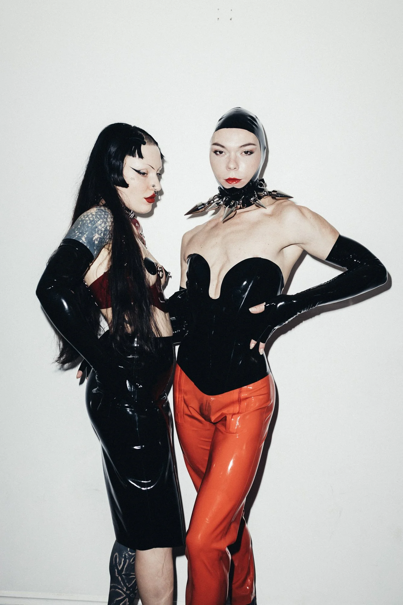 Two women with bold fashion style posing against a plain white wall, wearing latex outfits and striking confident poses.