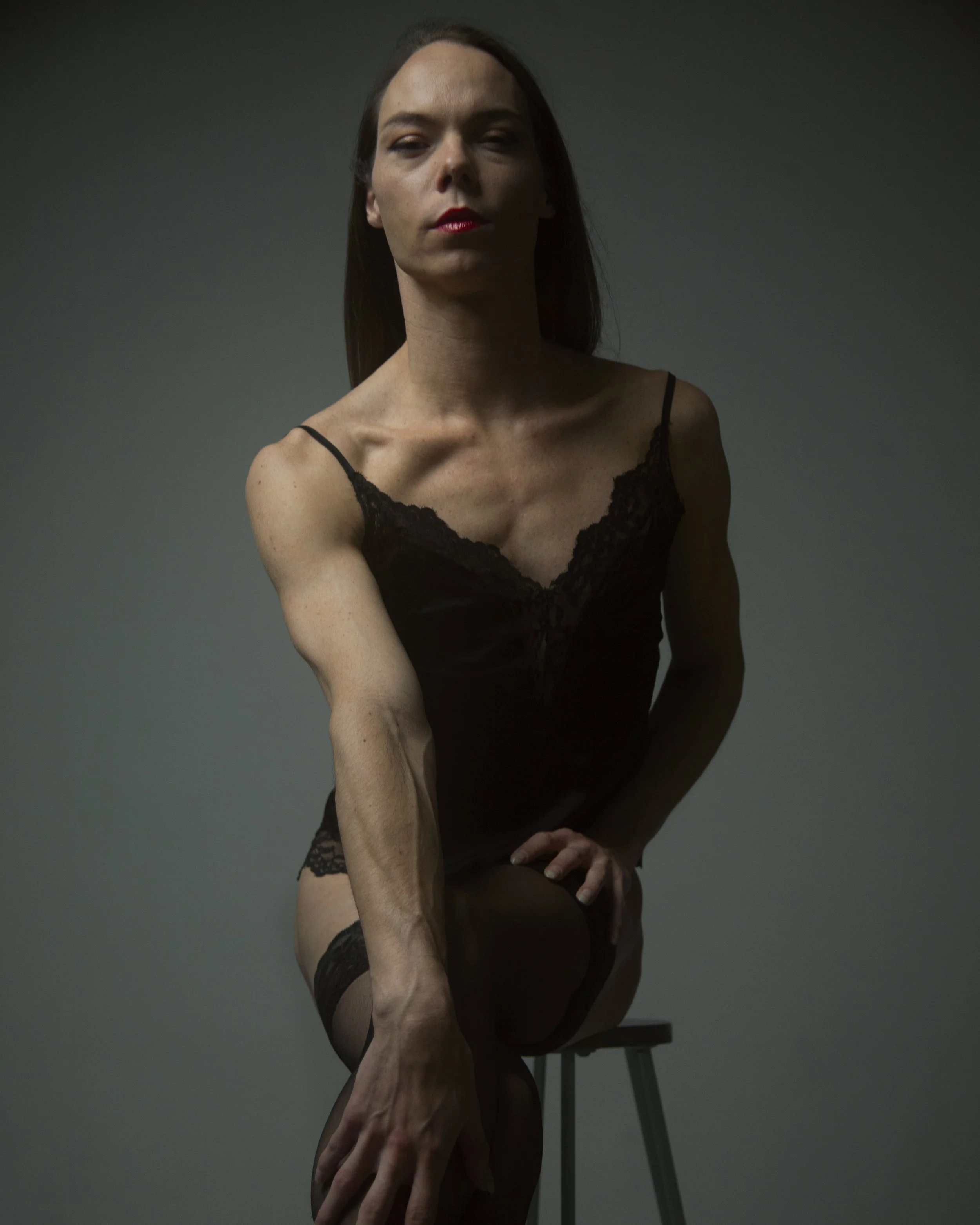 A woman with long dark hair, wearing black lace lingerie, sitting on a stool against a plain dark background, looking directly at the camera with a serious expression.