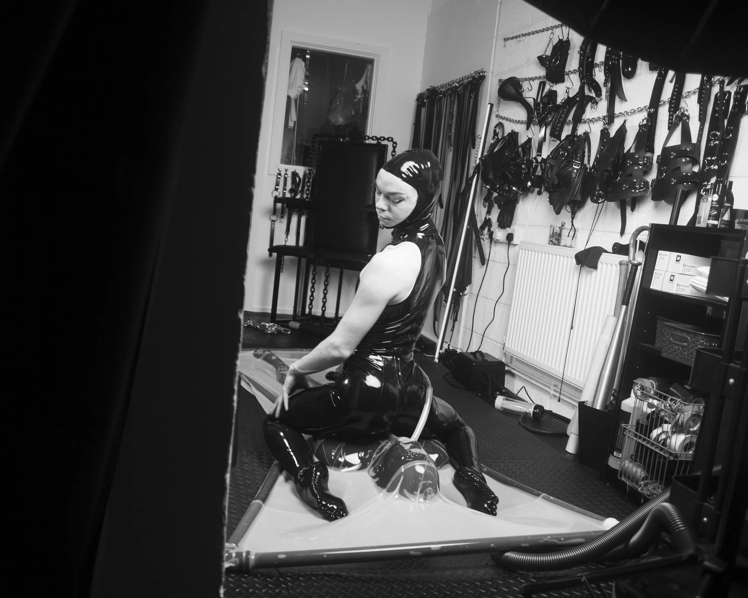 Black and white photo of a person sitting on a reflective surface, wearing a shiny, latex outfit and hood, inside a room with chains and leather accessories hanging on the walls.