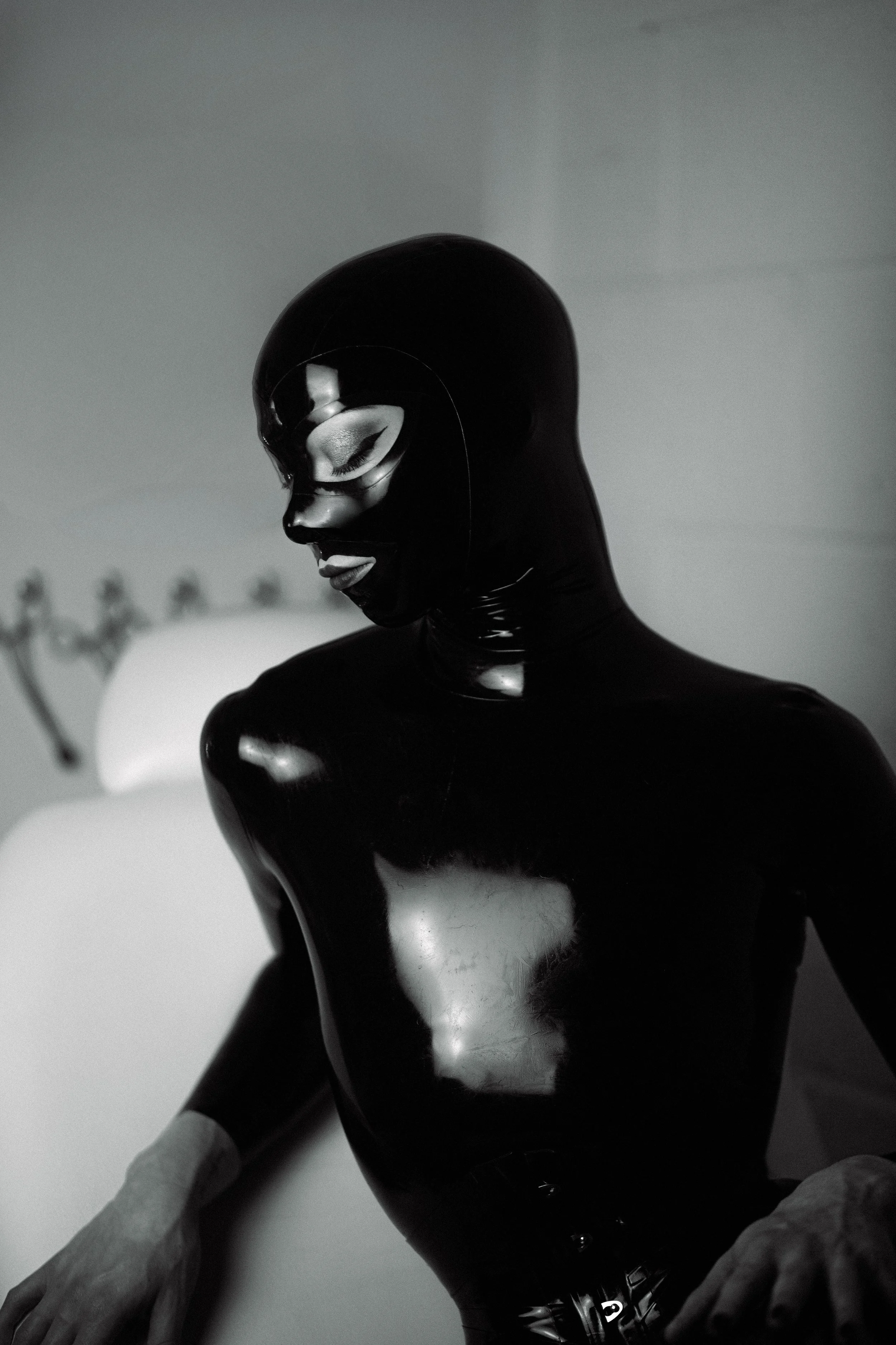 A person wearing a black, shiny latex bodysuit and a black mask with a face design, with closed eyes, and silver eye makeup.