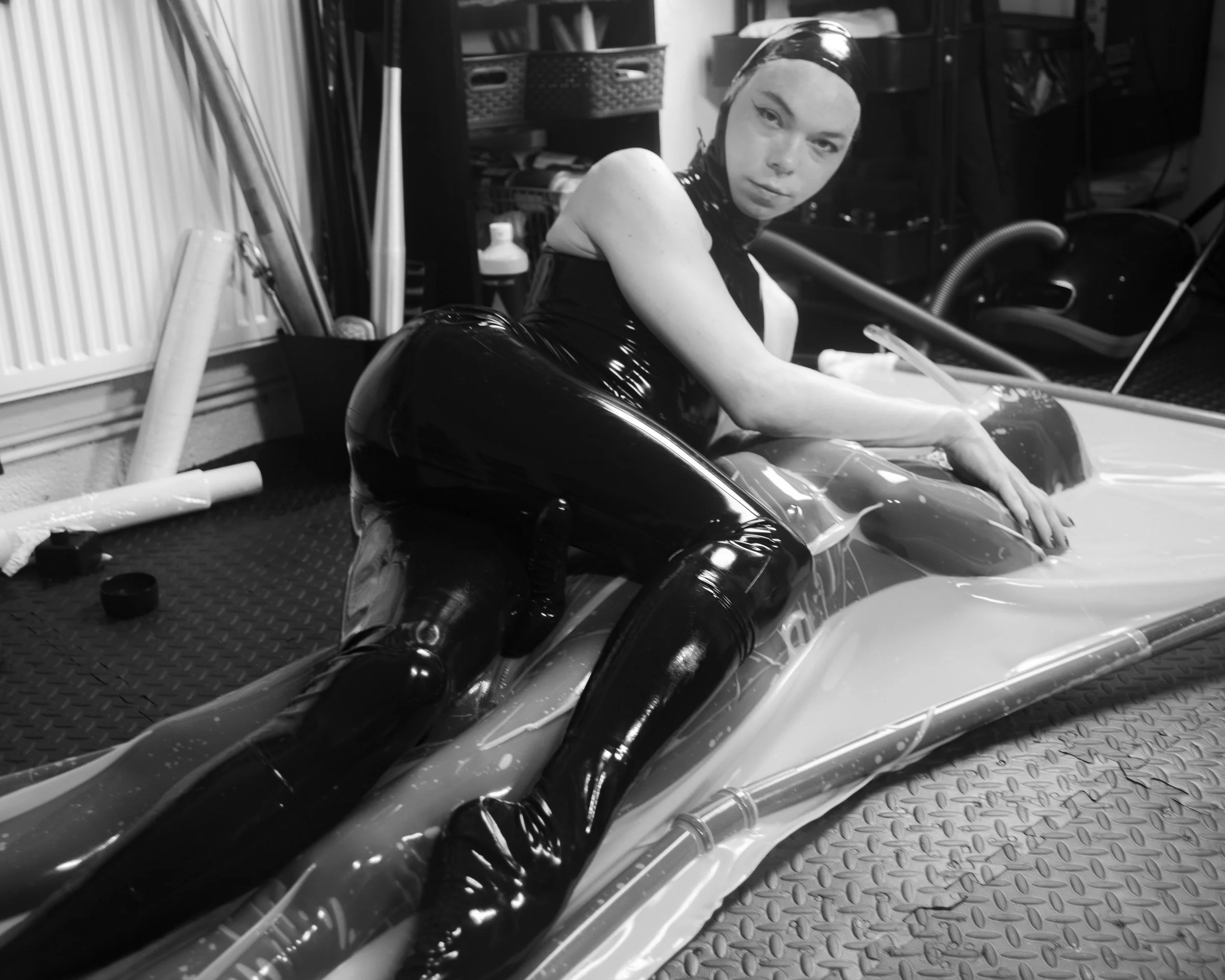 Black and white photo of a person with short hair, wearing a sleeveless top, sitting on the floor next to a shiny motorcycle in a garage or workshop.