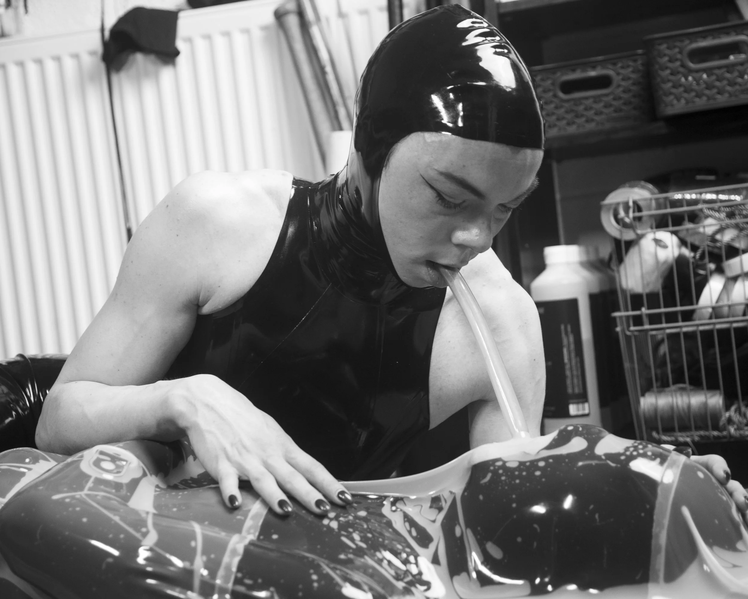 A woman wearing a shiny black latex mask and a sleeveless top, with her eyes closed, appears to be playing a wind instrument or pipe during a performance or practice, with a cluttered background of shelves and containers.