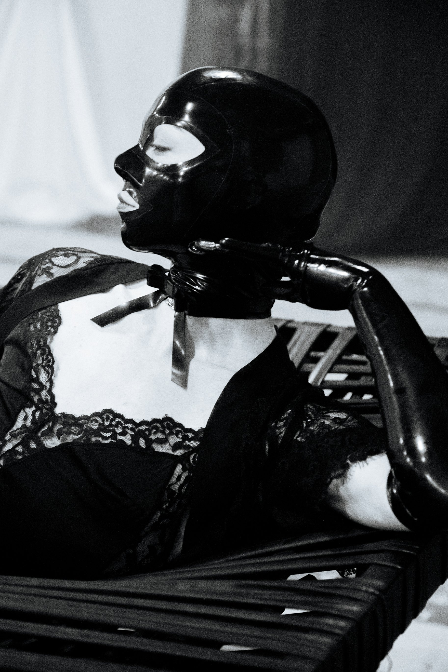 A woman with a painted face wearing a shiny black mask and lace clothing, sitting on a black bench with her hand resting on her neck, in a black and white photograph.