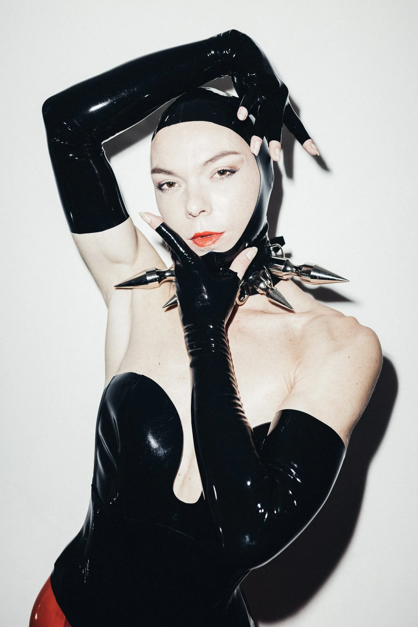 A person wearing black latex gloves, a black latex head covering, a spiked choker, and a black latex outfit poses with one arm raised and the other hand touching their face against a white background.
