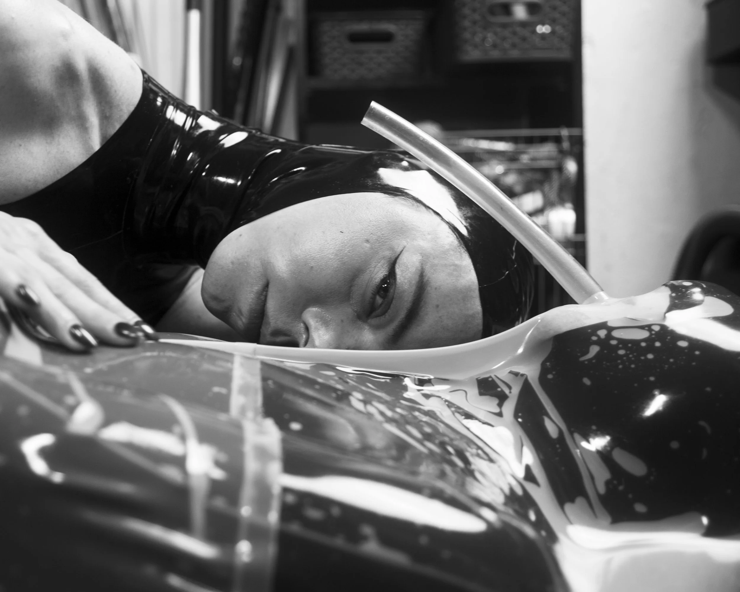 A person wearing a shiny black latex glove is drinking from a straw, with their face close to liquids in a bowl or container, in a kitchen or similar setting.