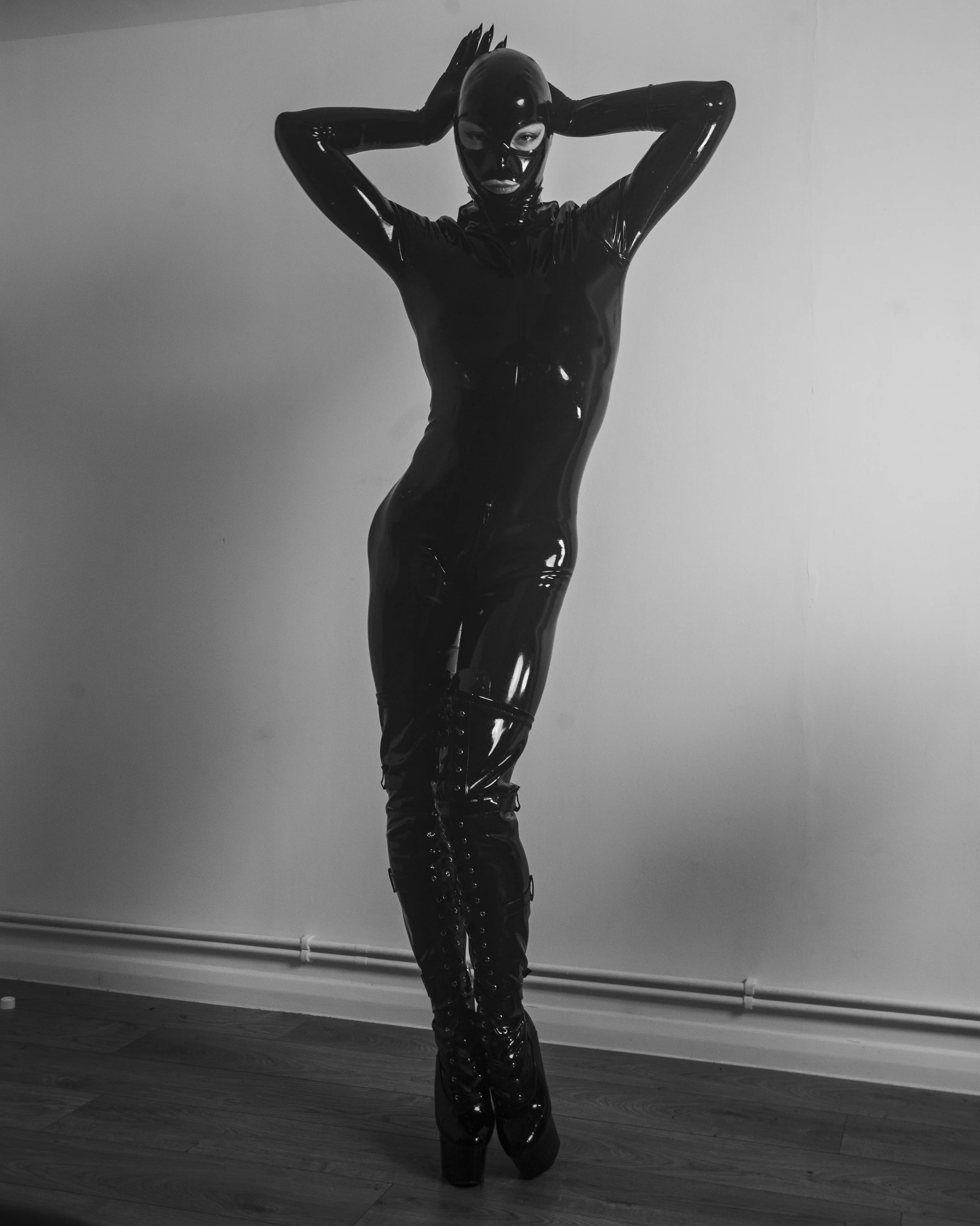 A person dressed in a shiny black latex bodysuit and high boots, wearing a black latex mask, standing with hands behind their head against a plain wall.