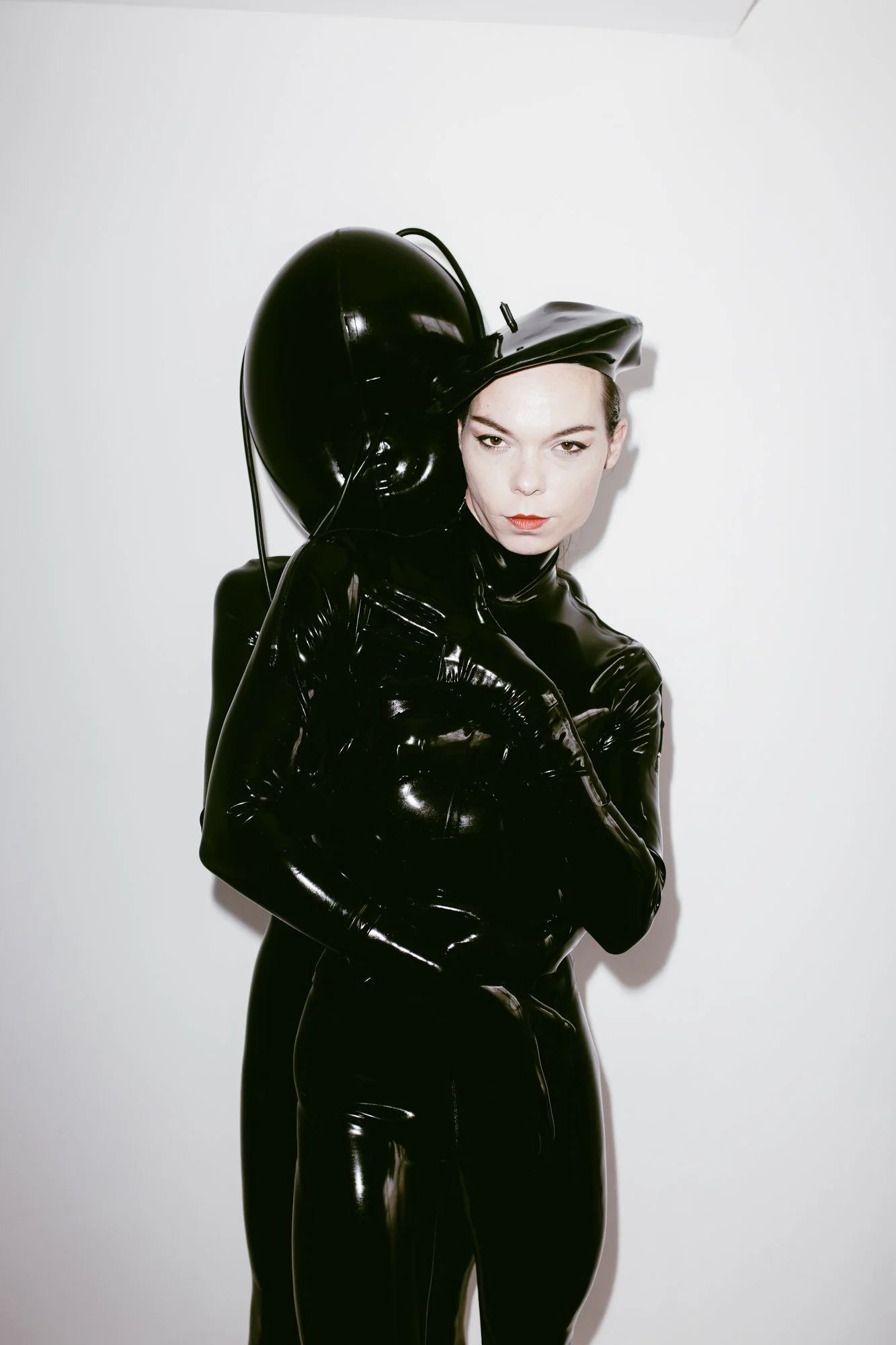 A woman dressed in a shiny black latex outfit, wearing a matching helmet or hat, posing against a plain white background.