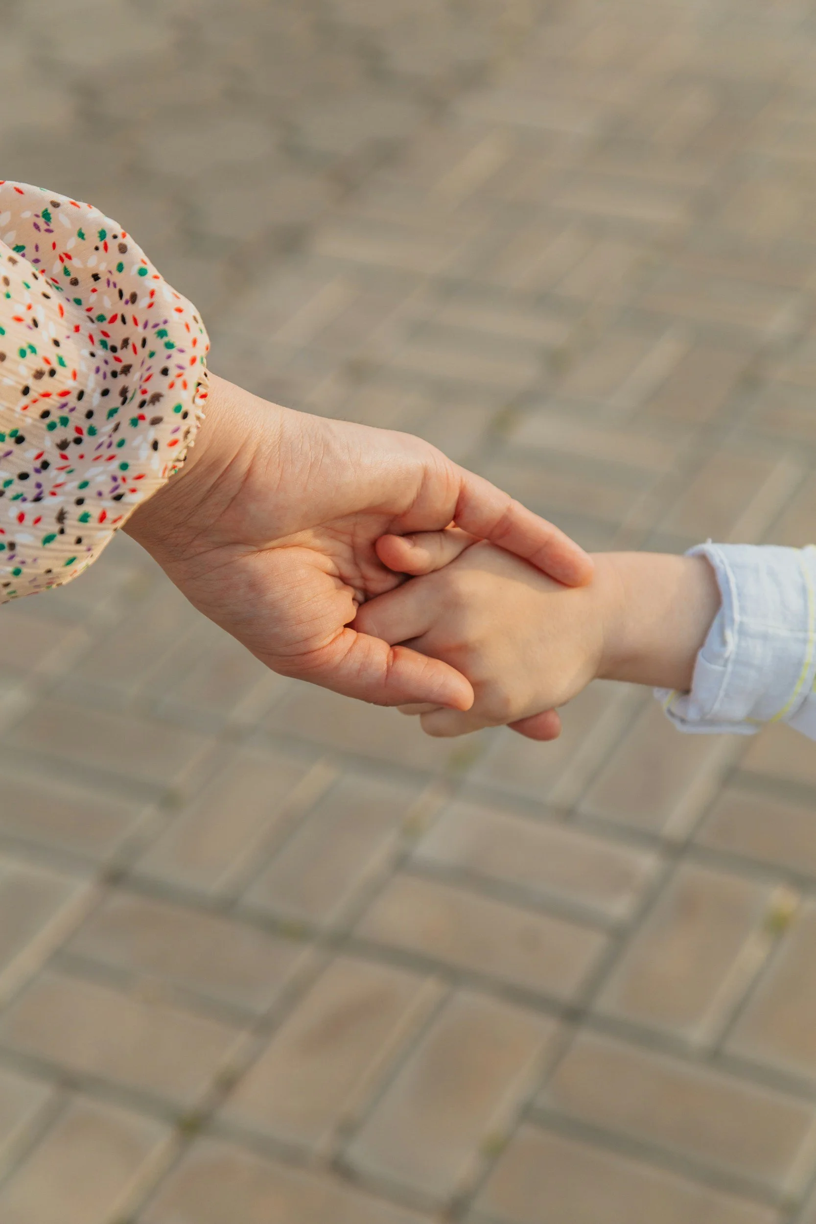 An adult's hand holding a child's hand, both hands are clasped with the child's hand on top, on a brick pavement background.
