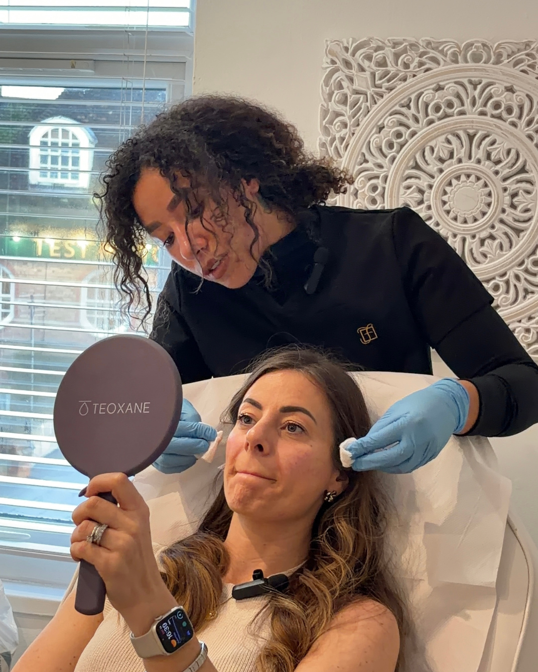 A woman lying in a bed holding a mirror with the word 'TEOXANE' on it. A beauty professional is applying a treatment to her face in a clinical setting, wearing blue gloves.