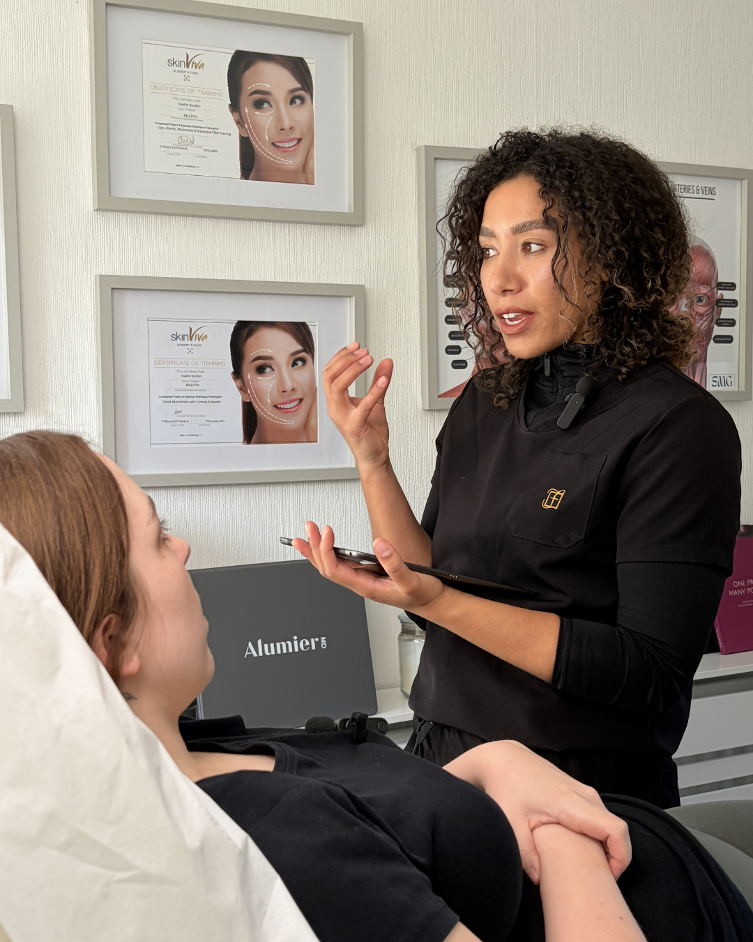 A woman with curly hair is talking to a patient lying on a medical bed, with medical posters on the wall behind her, including certificates and diagrams of the face.