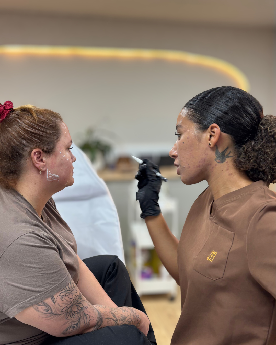 A tattoo artist is applying a tattoo to a woman sitting with her eyes closed in a studio.