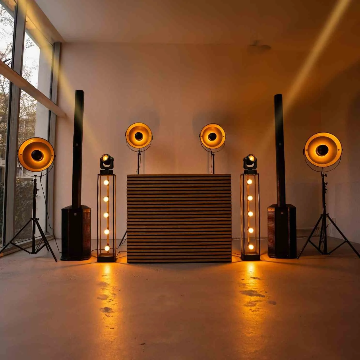 Indoor DJ setup with four orange speakers, two tall light stands with bulbs, and a black striped DJ booth, illuminated in warm orange lighting.