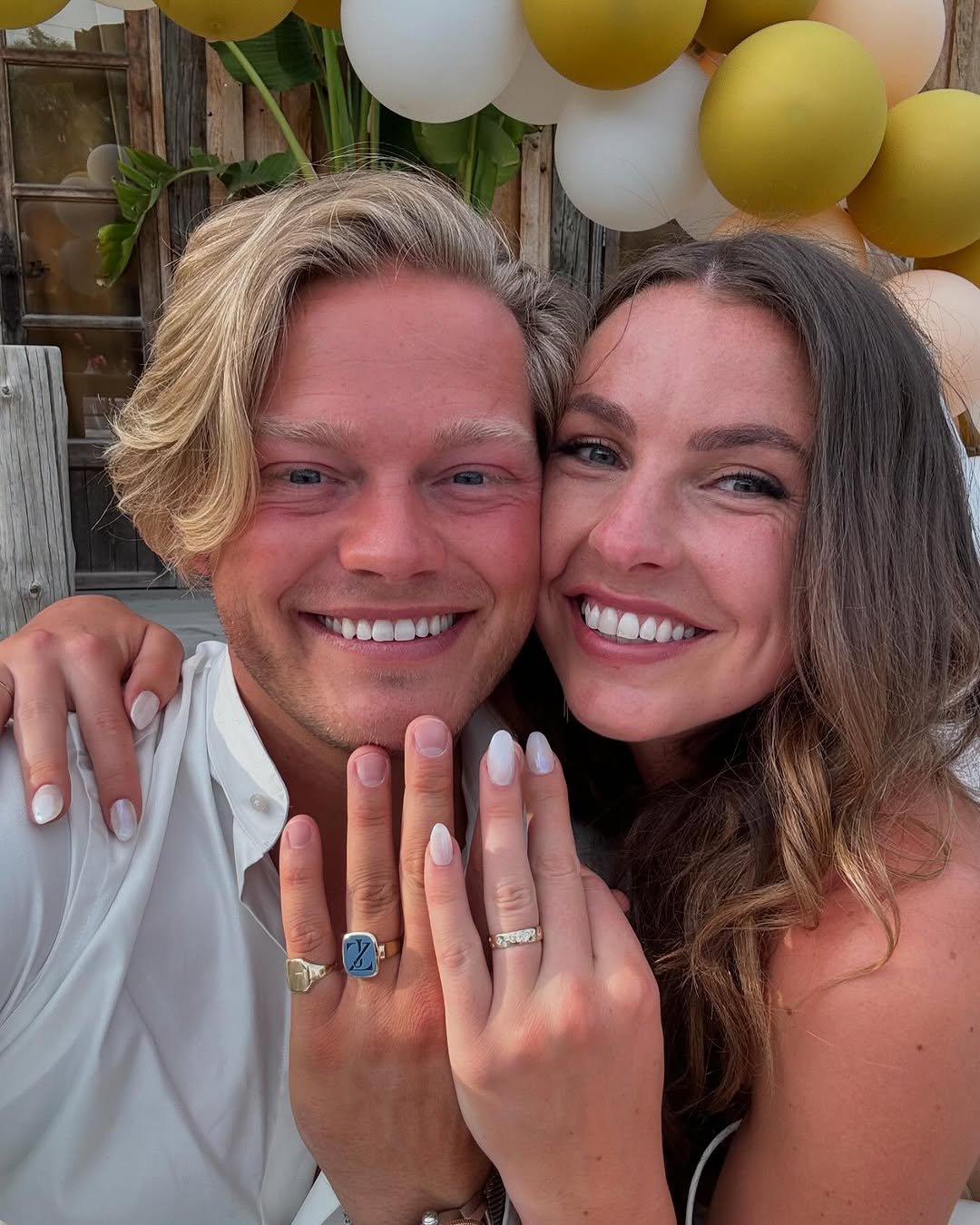 A happy couple smiling and showing their rings, celebrating an engagement or wedding, with balloons and a wooden backdrop.