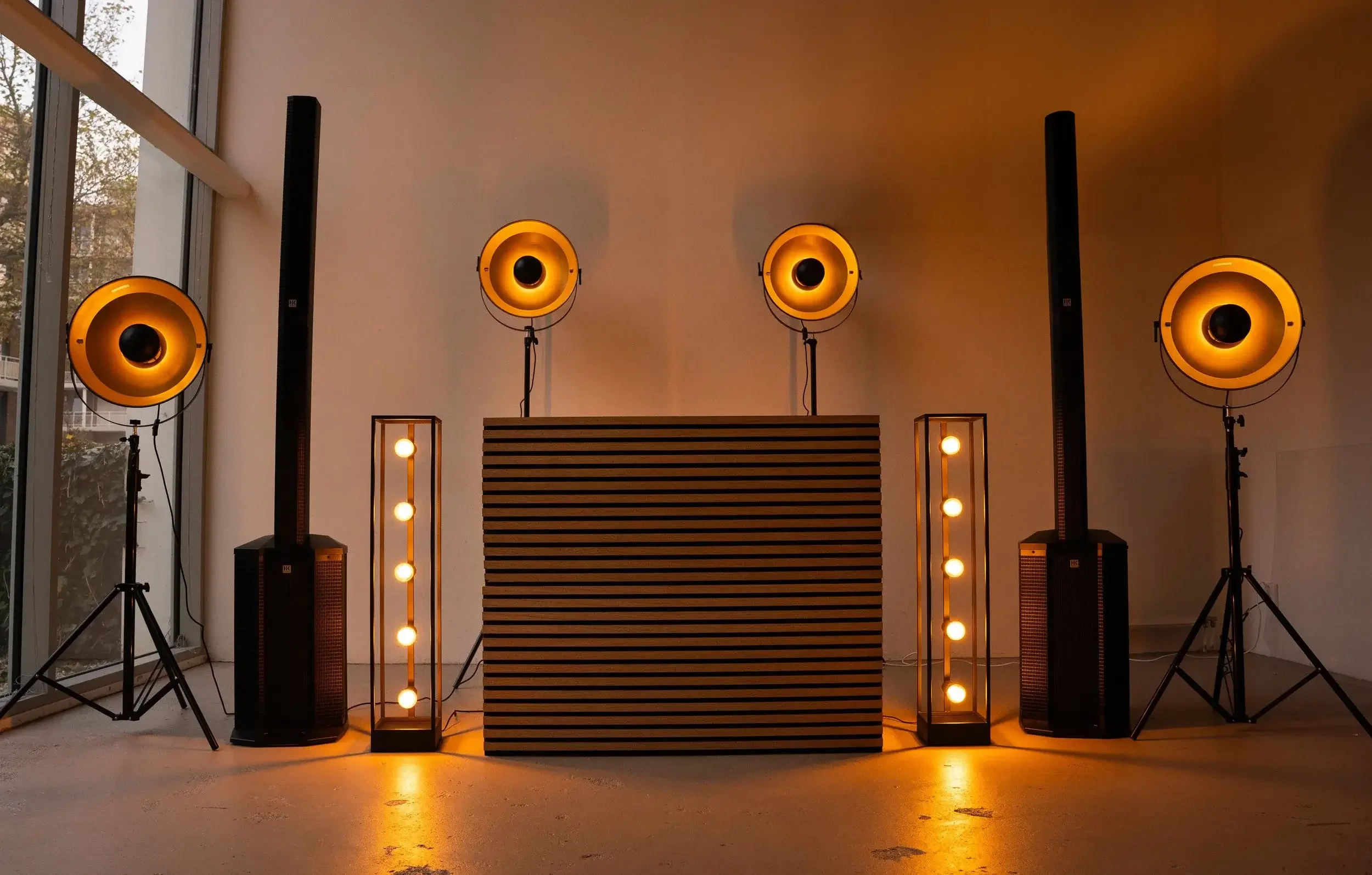 Music or event setup with large yellow stage lights, vertical speakers, illuminated floor lamps, and a wooden textured wall in a bright room with large windows.