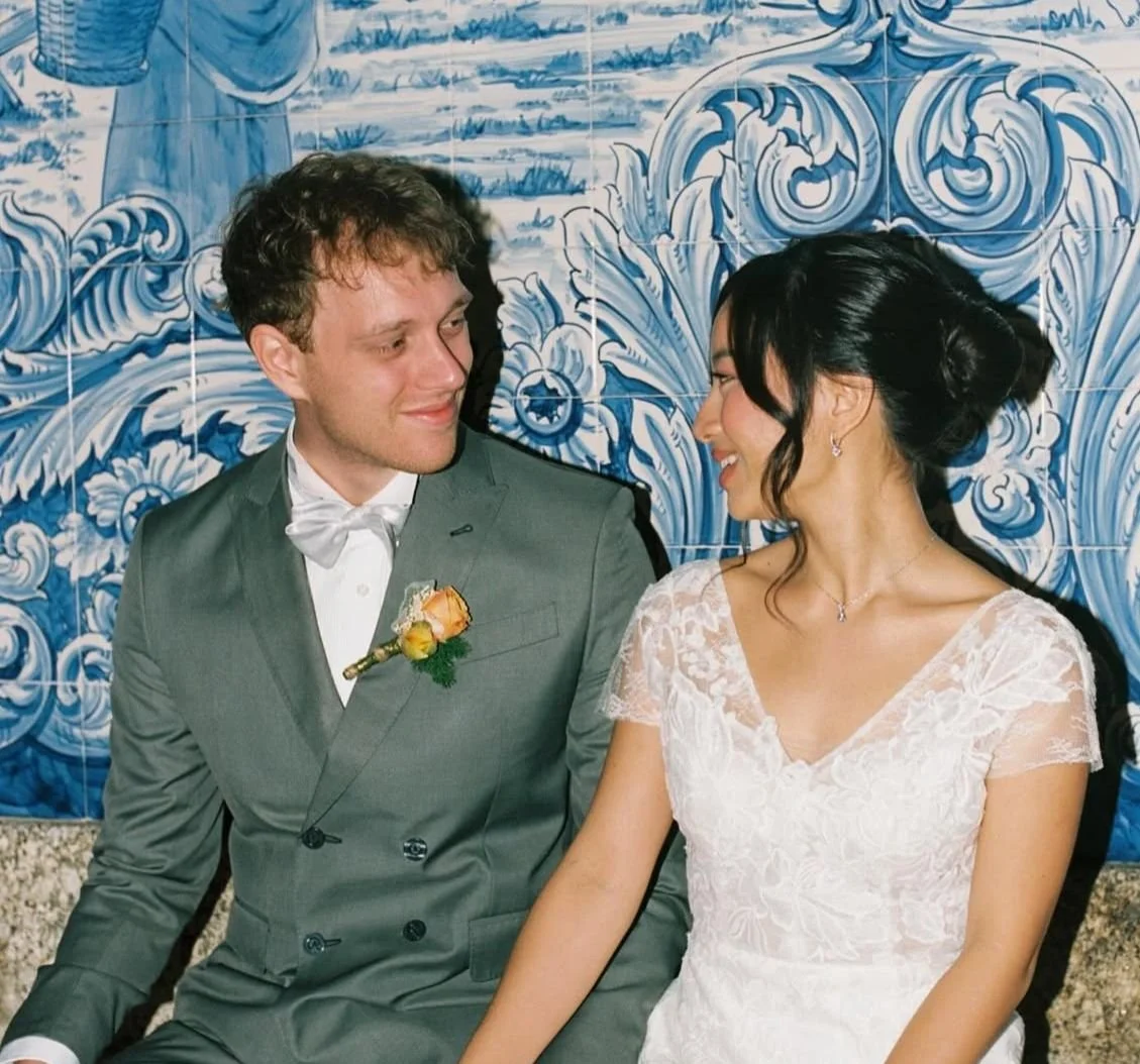 A bride and groom sitting close together, smiling at each other, with a blue and white decorative tiled wall in the background.