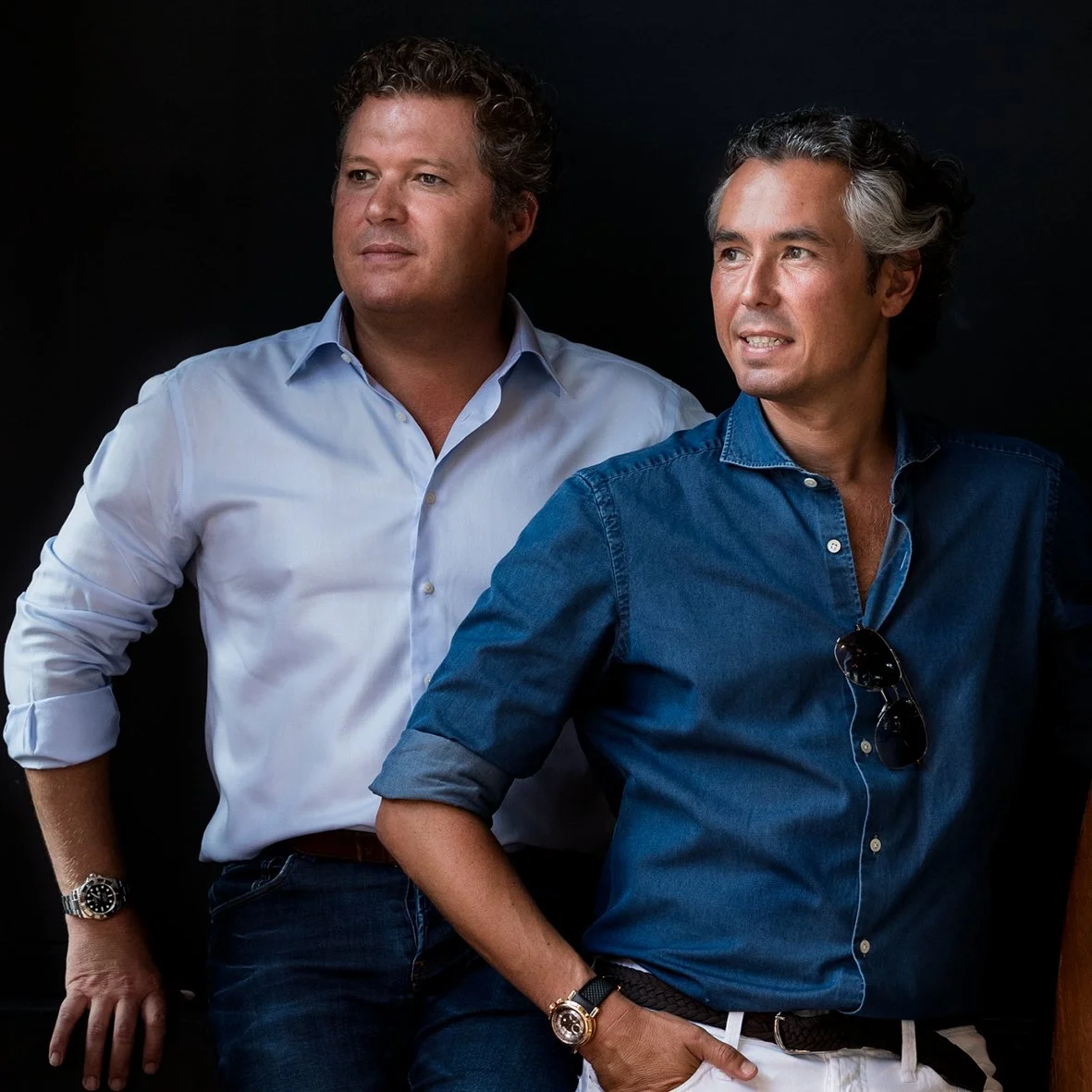 Two men with gray hair and light skin, wearing casual shirts, standing against a dark background. The man on the left has curly hair, a light blue shirt, and a watch, while the man on the right has wavy hair, a denim shirt with sunglasses hanging from the collar, and a watch.