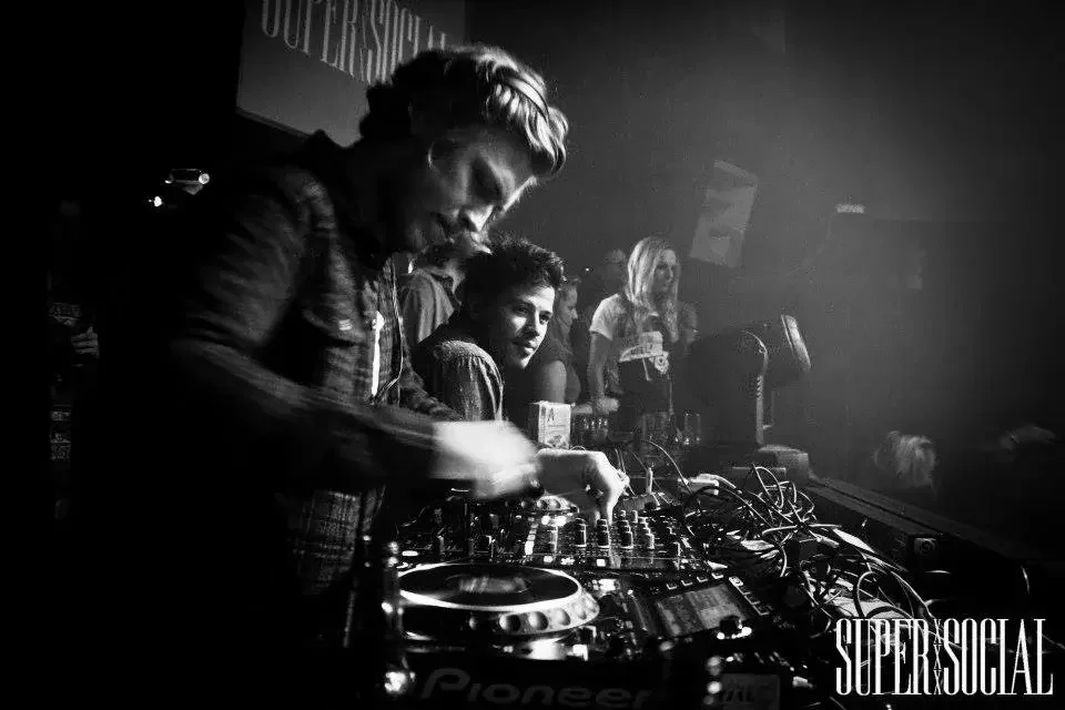 A DJ with short, light-colored hair plays music at a club or party with multiple people dancing and socializing in the background.