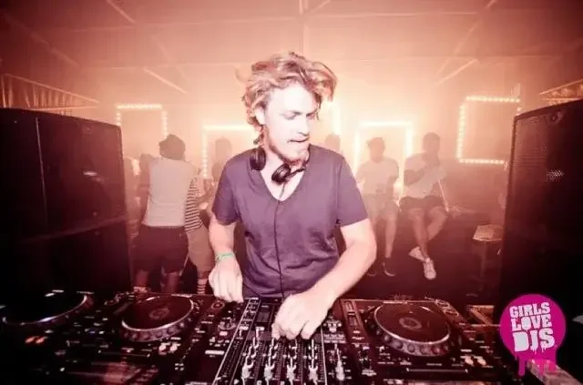 A DJ with curly hair and headphones around his neck plays music at a nightclub with purple lighting, while people dance in the background. A pink logo in the lower right corner reads 'Girls Love DJs'.