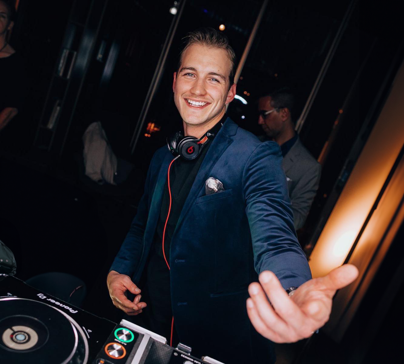 A smiling young man in a navy blue blazer with a pocket square, wearing headphones around his neck, standing next to a DJ controller at an indoor event.