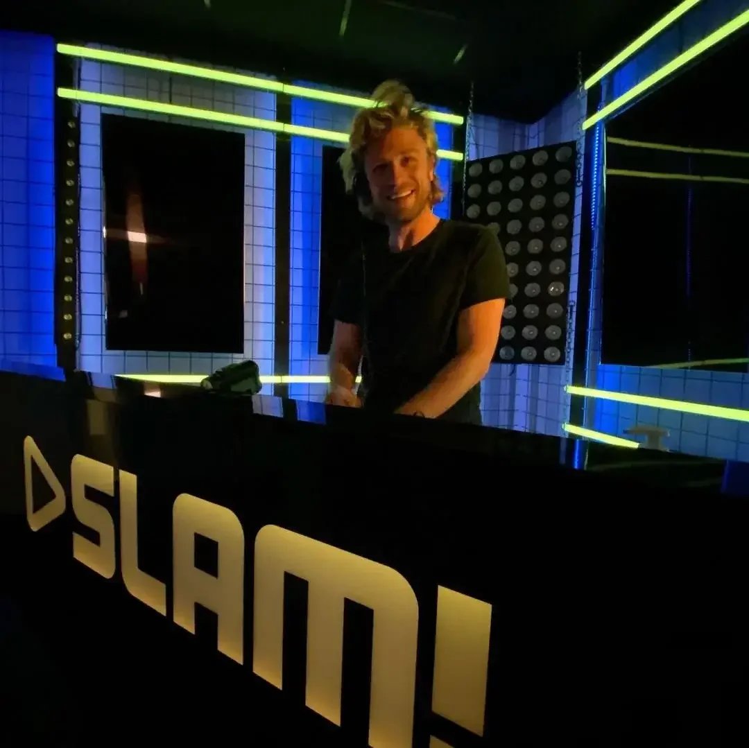 A smiling DJ wearing a black shirt, standing behind a DJ booth with a sign that says 'SLAND,' inside a club with blue tiled walls and neon yellow light strips.
