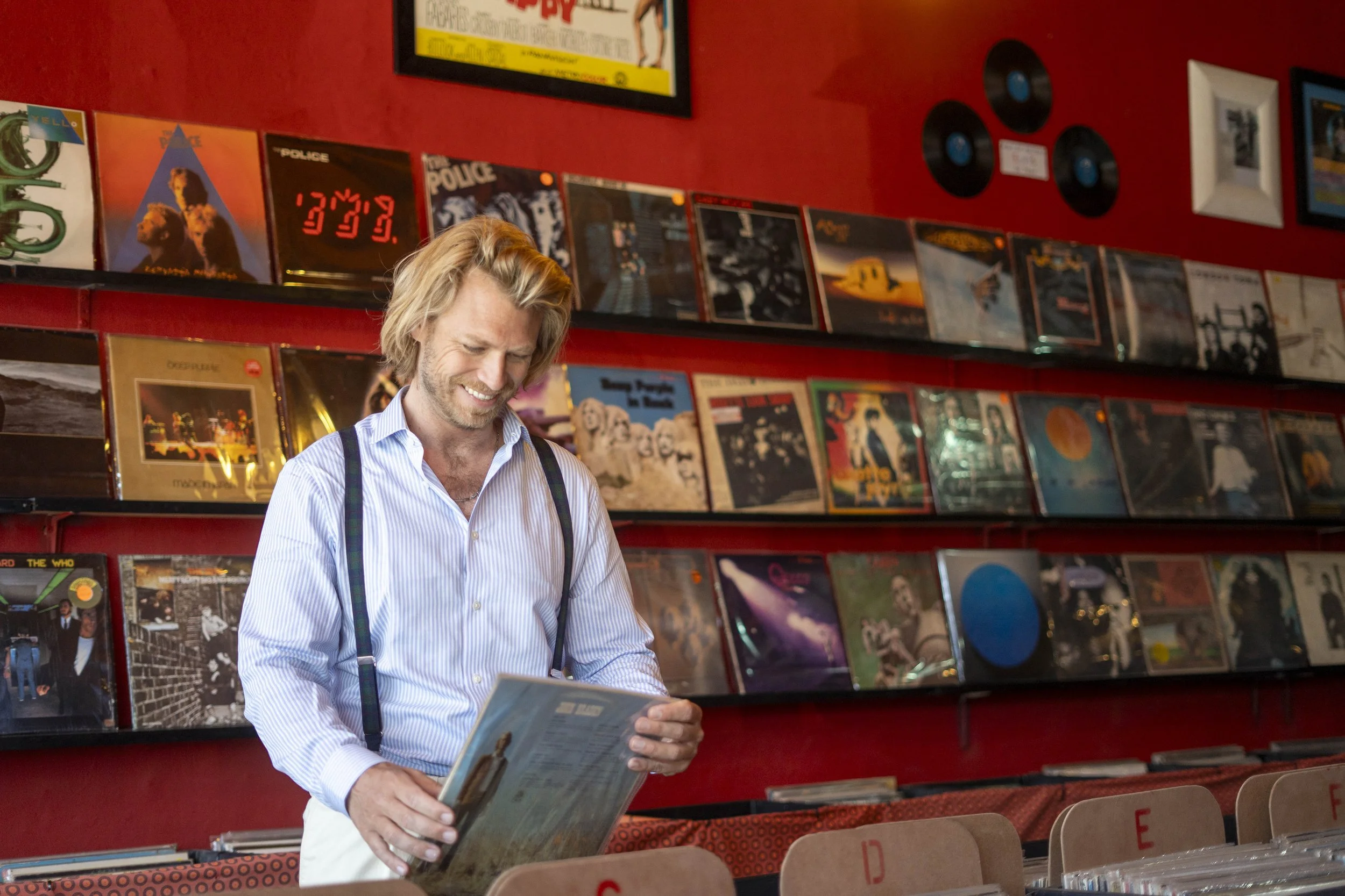 A man with blonde hair and a light blue striped shirt with suspenders browsing a vinyl record at a record store with red walls and shelves filled with albums.