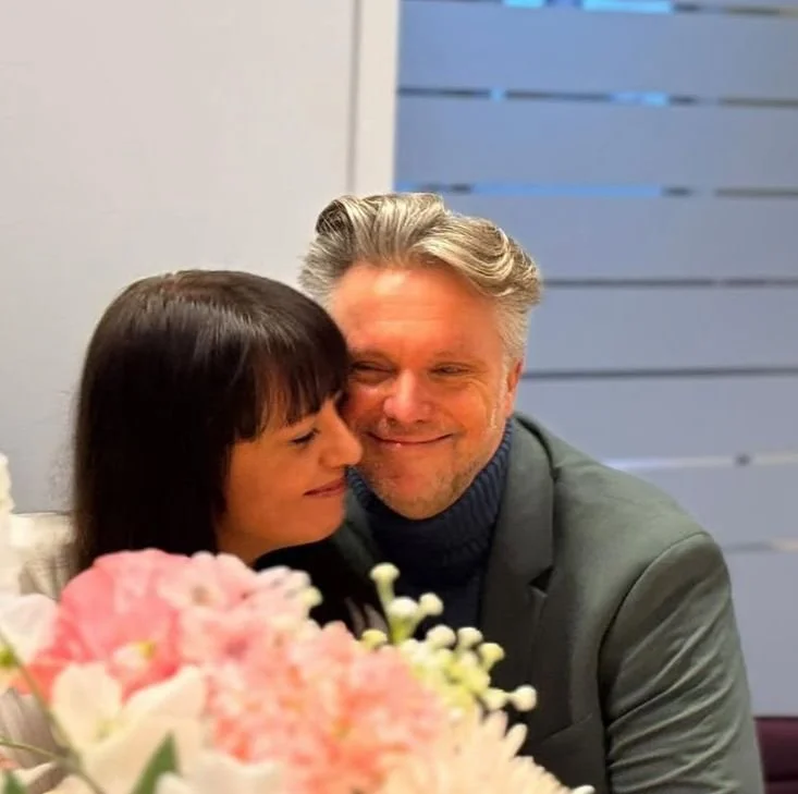 A man and woman sitting close together, smiling, with pink and white flowers in front of them. The woman has dark hair and the man has gray hair.