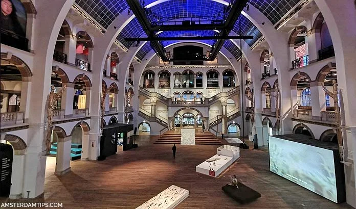 Interior of a large, architectural building with multiple arches, balconies, and a glass ceiling, featuring exhibition displays and seating areas.