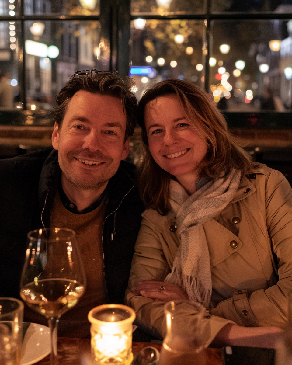 A smiling couple sitting at a restaurant table at night, with blurred city lights and windows behind them. There is a glass of white wine and a lit candle on the table.