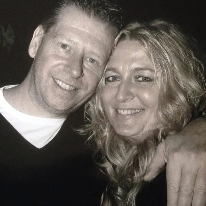 A smiling man and woman taking a close-up selfie together. The man has short hair and is wearing a dark sweater with a white shirt underneath. The woman has curly blonde hair and is smiling with her eyes slightly squinted. The background is dark and blurry.