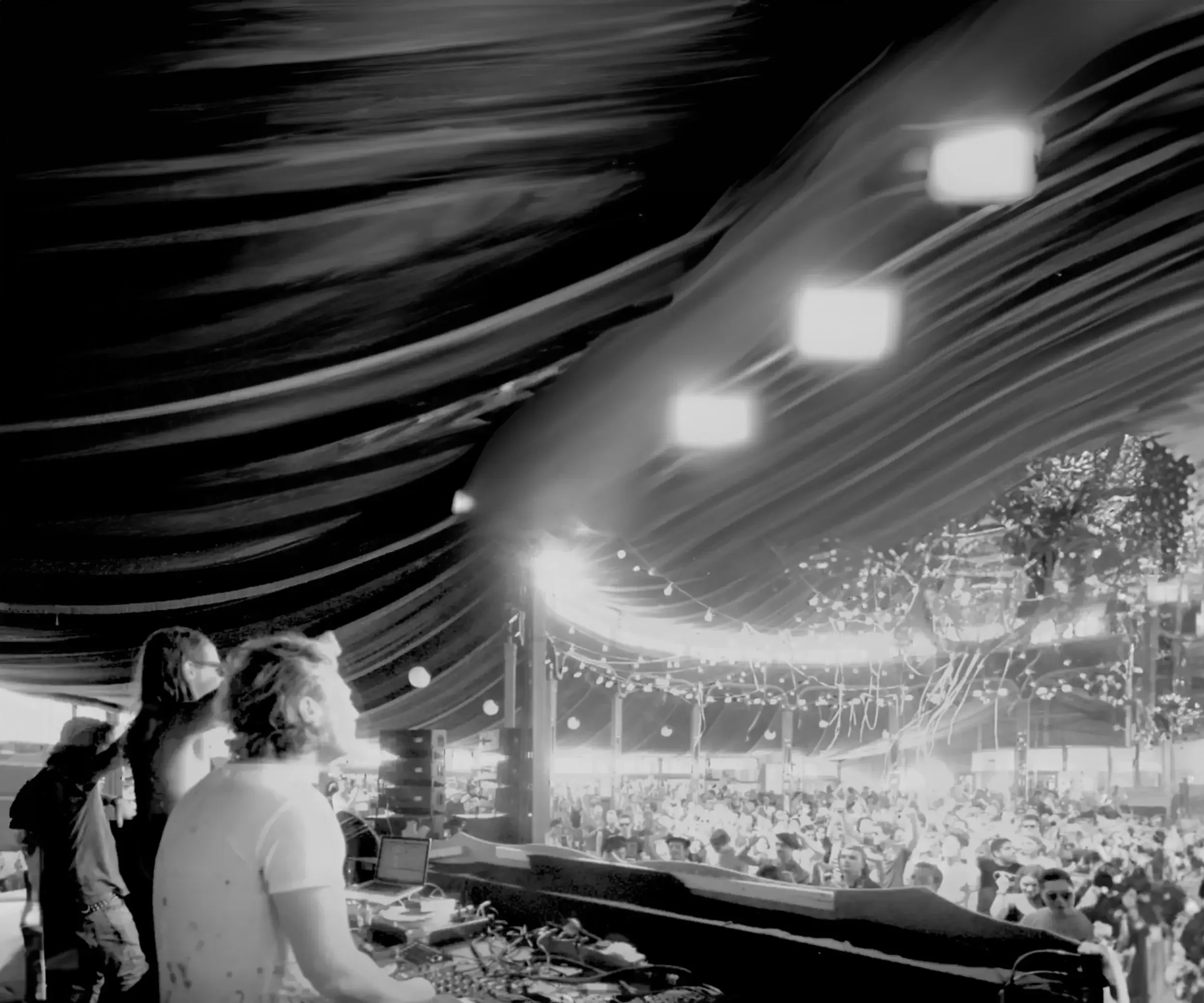 People DJing at a crowded indoor music event with string lights and fabric drapes on the ceiling.