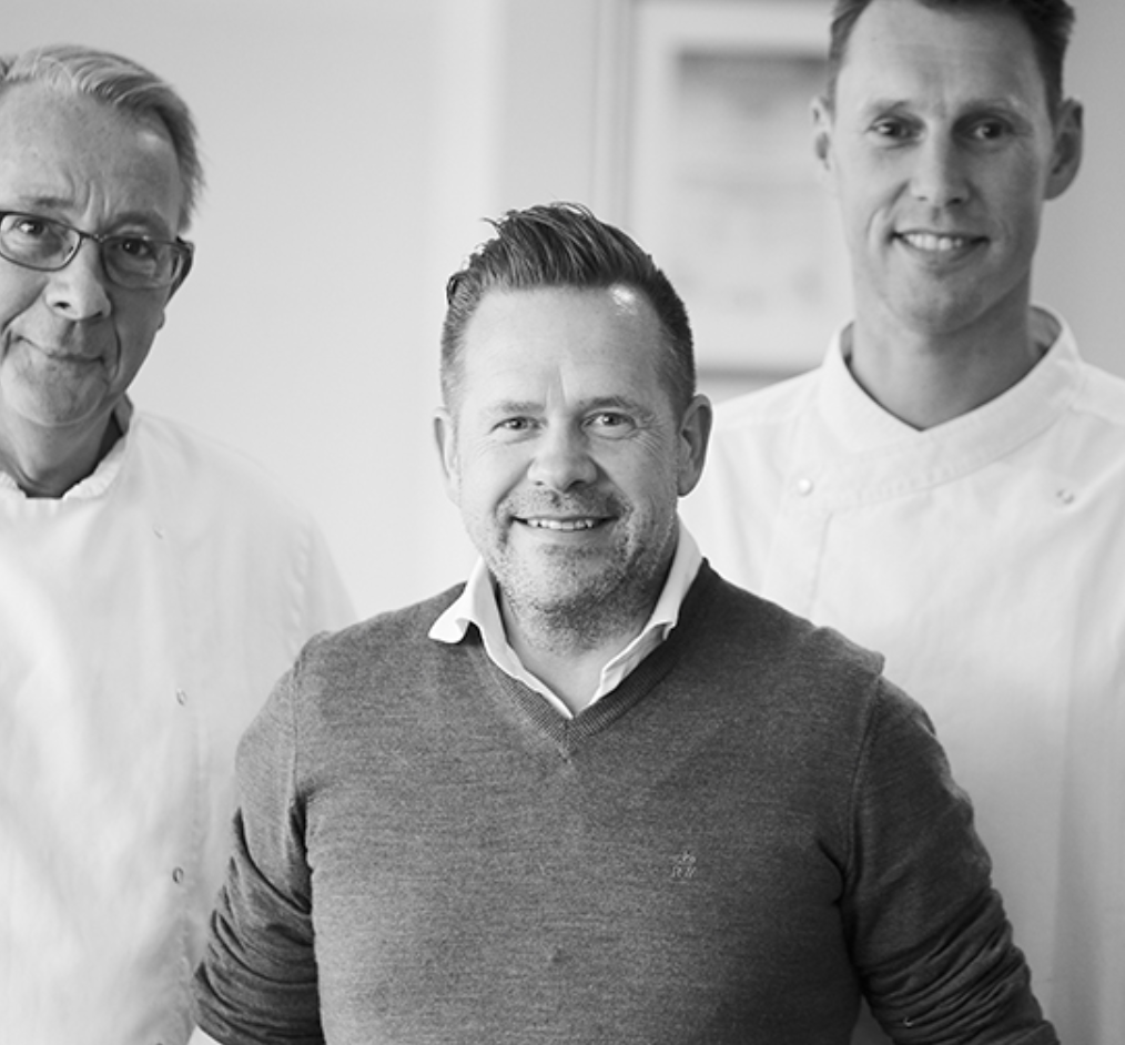Black and white photo of three men, two dressed as chefs, standing in a kitchen. The man in the center has short hair, a beard, and is wearing a sweater over a collared shirt. The man on the left wears glasses and a chef's coat, and the man on the right has short hair and is also in a chef's coat.
