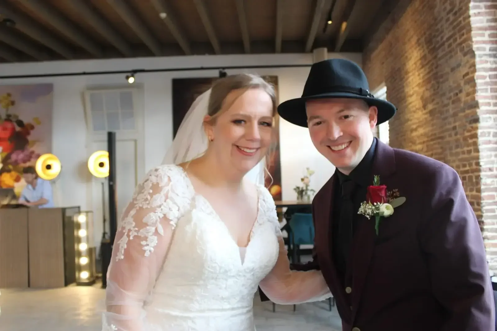 A bride and groom smiling together indoors at their wedding, in front of a brick wall. The bride is wearing a white lace wedding dress with sheer lace sleeves and a veil, while the groom is wearing a dark suit with a black fedora and a boutonnière.