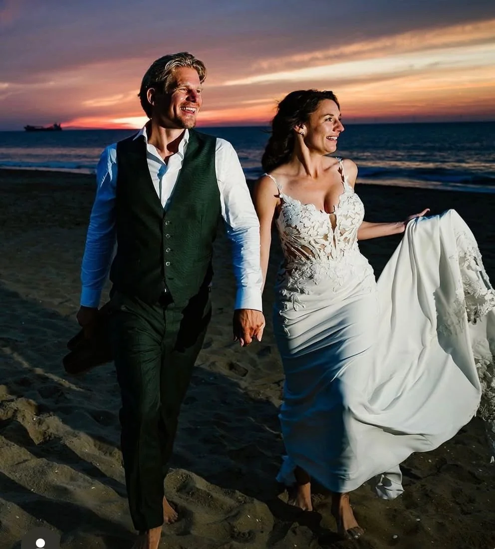 A couple in wedding attire walking on the beach at sunset, holding hands and smiling.