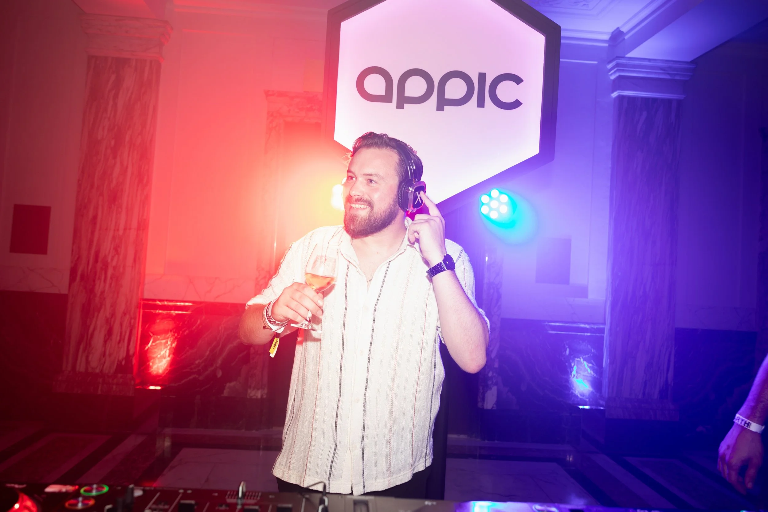 A man with a beard wearing a white shirt and headphones is holding a glass of wine while smiling. He is standing in front of a DJ setup at an event with colorful lighting. There is a large screen behind him displaying the word 'appic'.