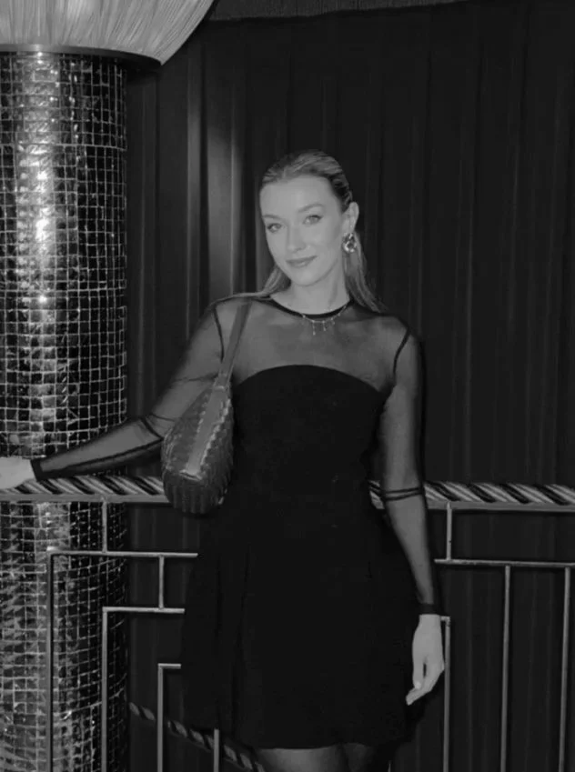 A young woman with light hair and a confident expression standing in front of red curtains and a gold railing, wearing a black dress with sheer long sleeves and carrying a red handbag.