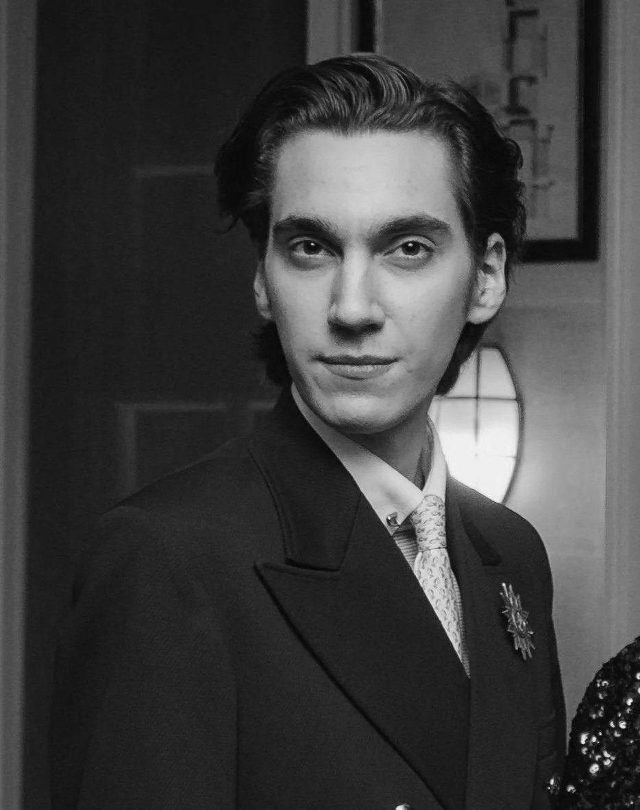 A young man with wavy brown hair wearing a dark suit, white shirt, and light-colored tie, standing indoors with warm lighting and framed pictures in the background.