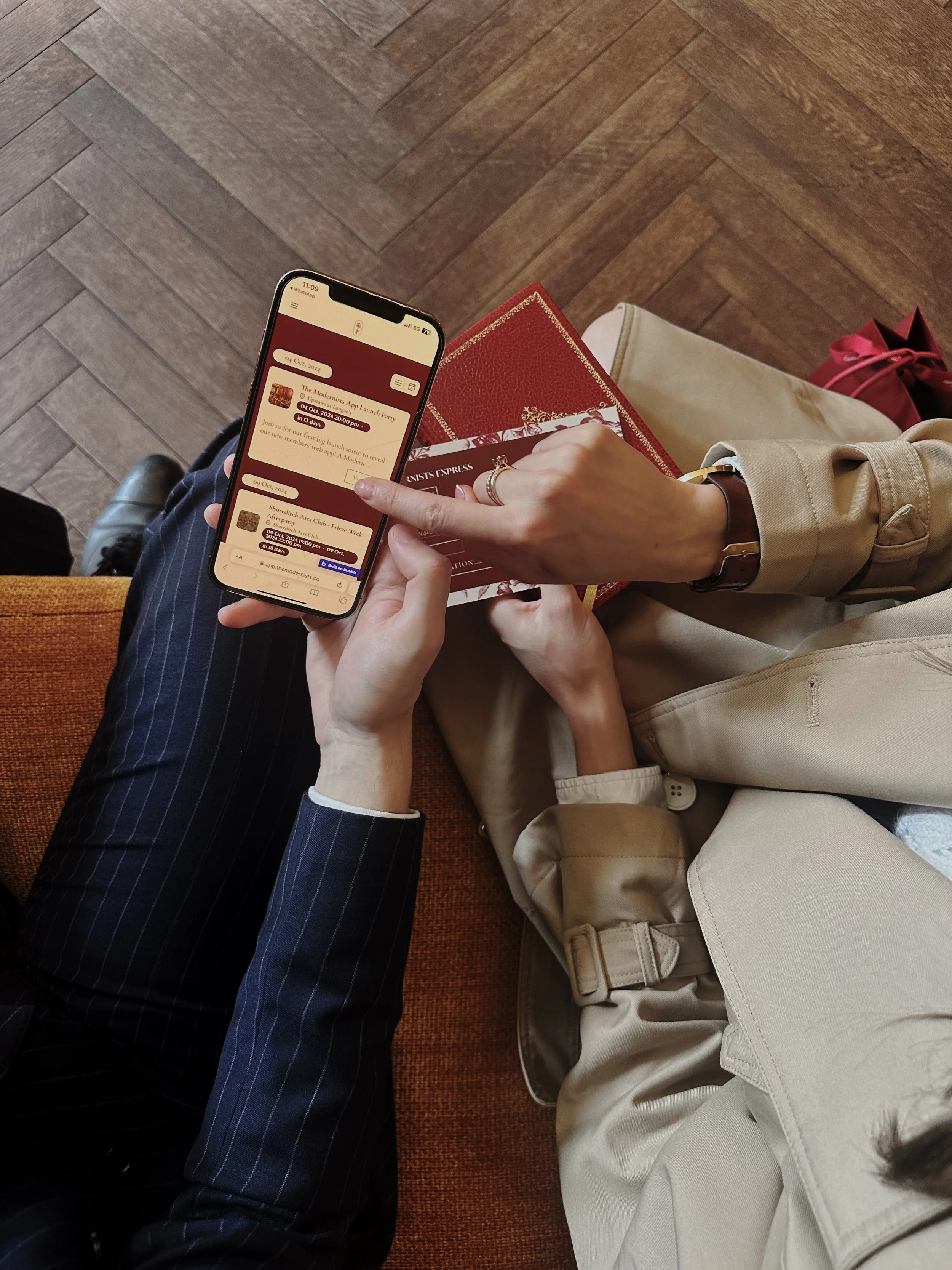 A person in a beige trench coat holding a phone and pointing at an app, sitting on a brown couch with another person in pinstripe pants. The person is also holding a red booklet or ticket. The setting is indoors with wooden flooring.