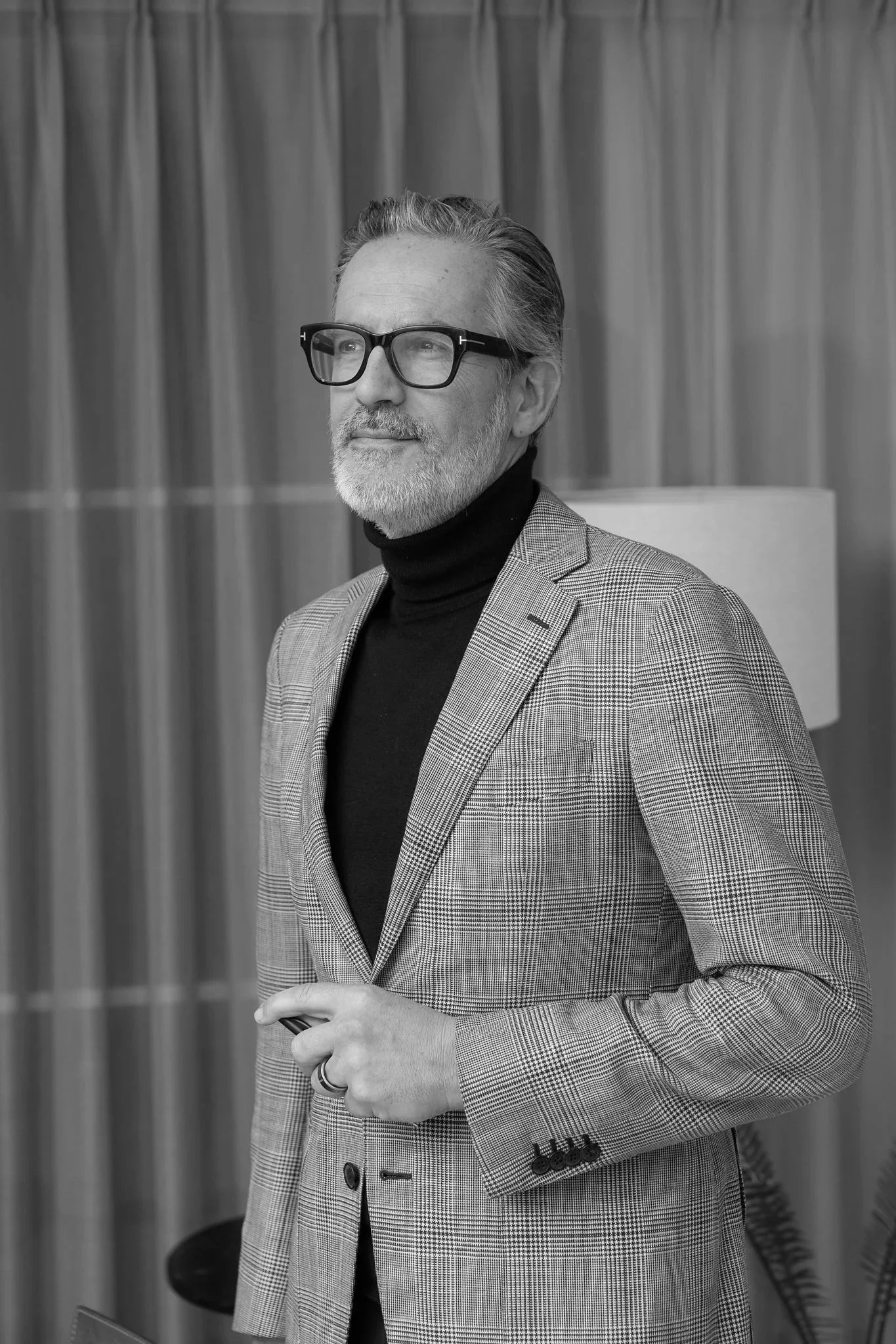 A distinguished older man with glasses, a beard, and gray hair, wearing a checkered blazer and a black turtleneck, standing indoors in front of curtains and a lamp.