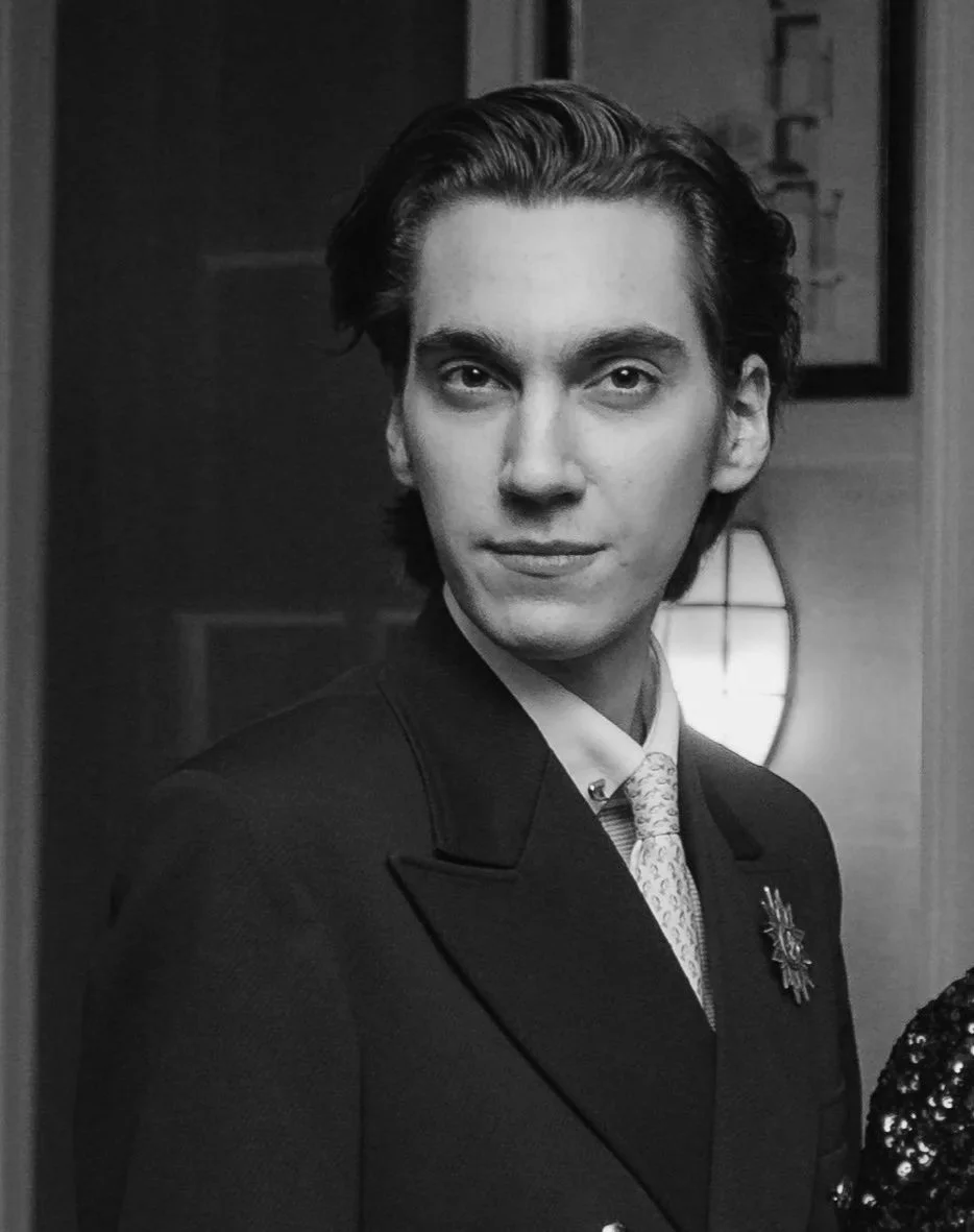 A young man with wavy brown hair wearing a dark suit, white shirt, and light-colored tie, standing indoors with warm lighting and framed pictures in the background.