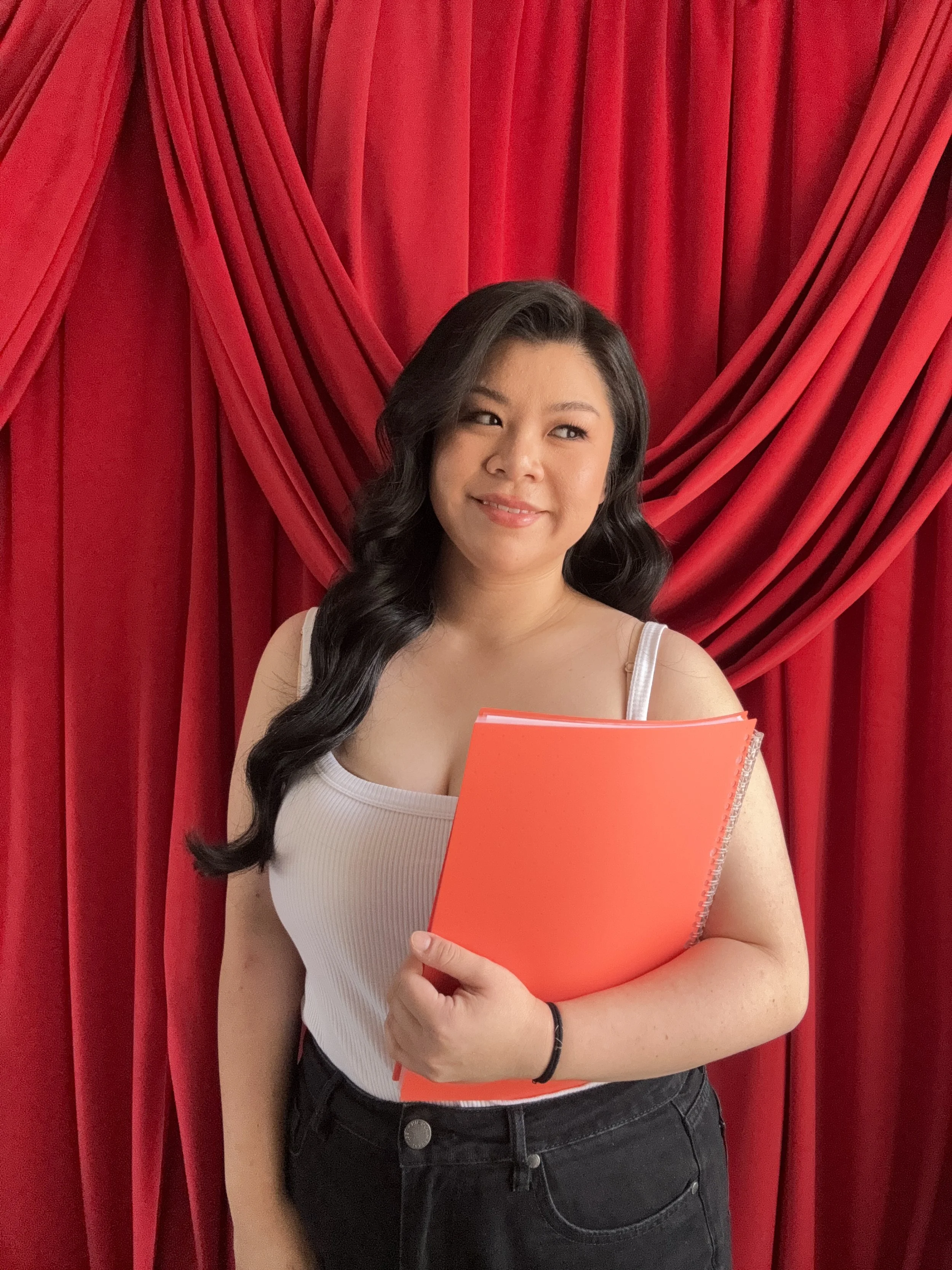 A young woman with long, wavy black hair standing in front of a red curtain, holding an orange notebook, wearing a white tank top and black jeans, smiling softly.