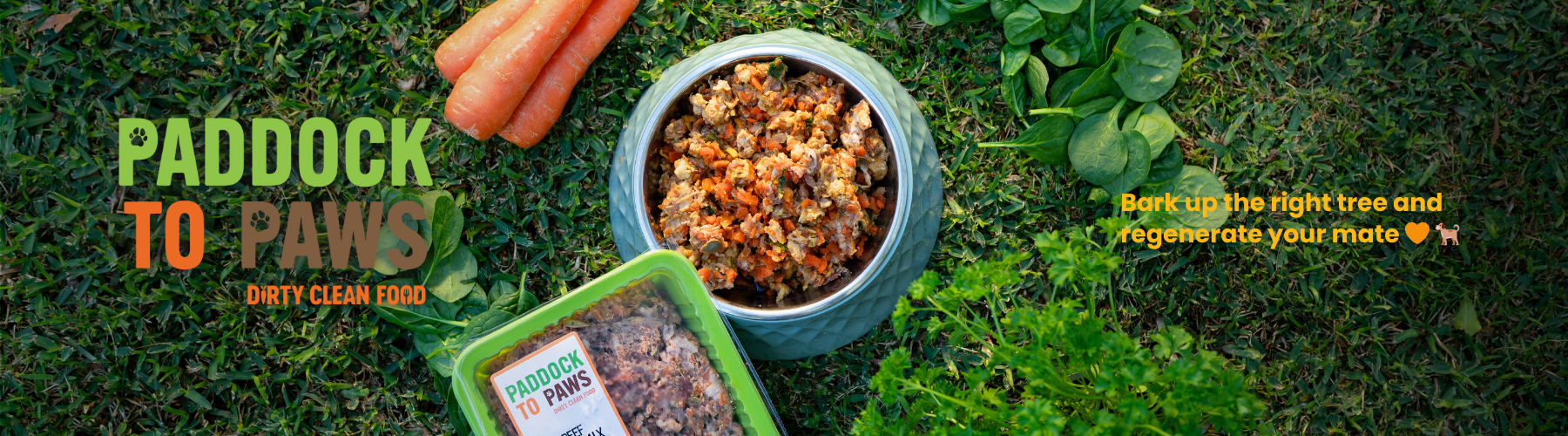 Dog food on a grassy ground with carrots, leafy greens and a container of ground meat labeled "Paddock to Paws" a slogan encouraging dog owners to feed natural food, with a caption "Bark up the right tree and regenerate your mate".