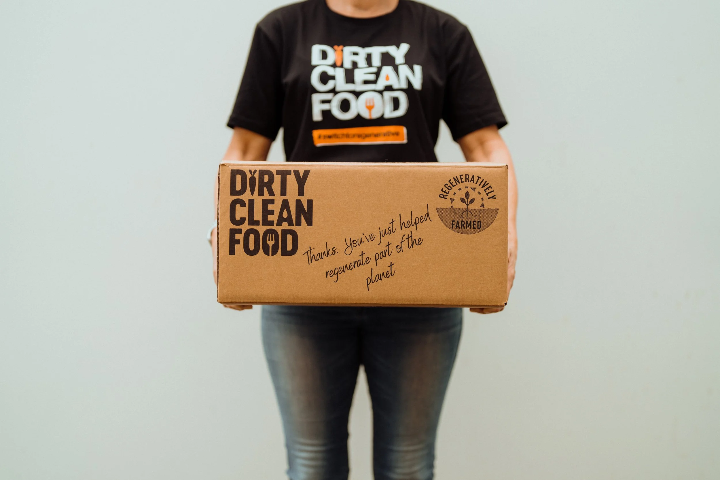 Person holding a cardboard box with 'Dirty Clean Food' branding and a message about regenerating part of the planet, wearing a black t-shirt with the same branding.