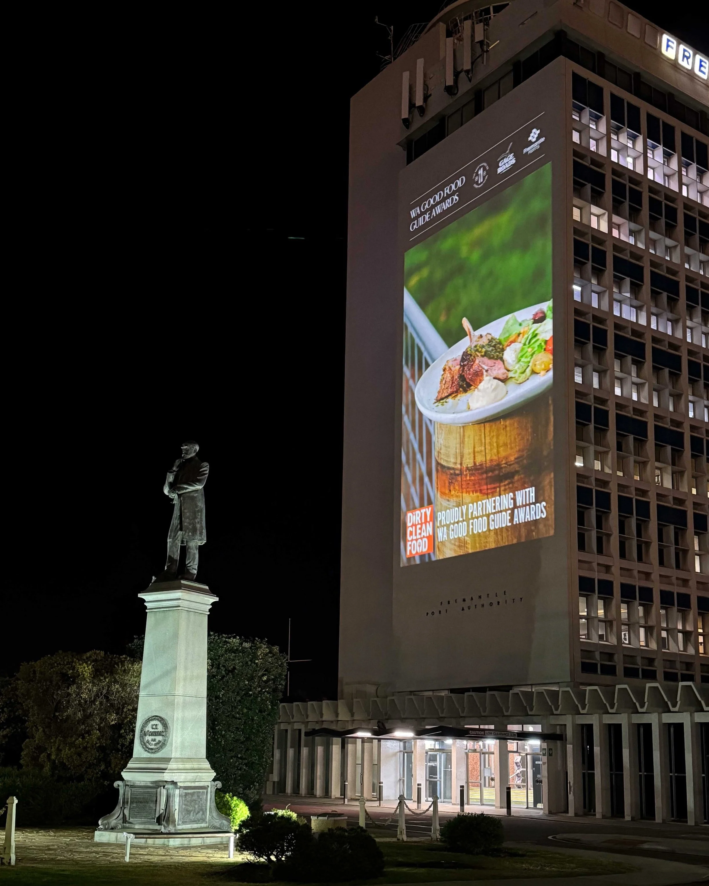 Nighttime view of a tall building with a large digital billboard displaying food, advertisements, and awards. To the left, there is a statue of a man on a pedestal, with trees and bushes below.