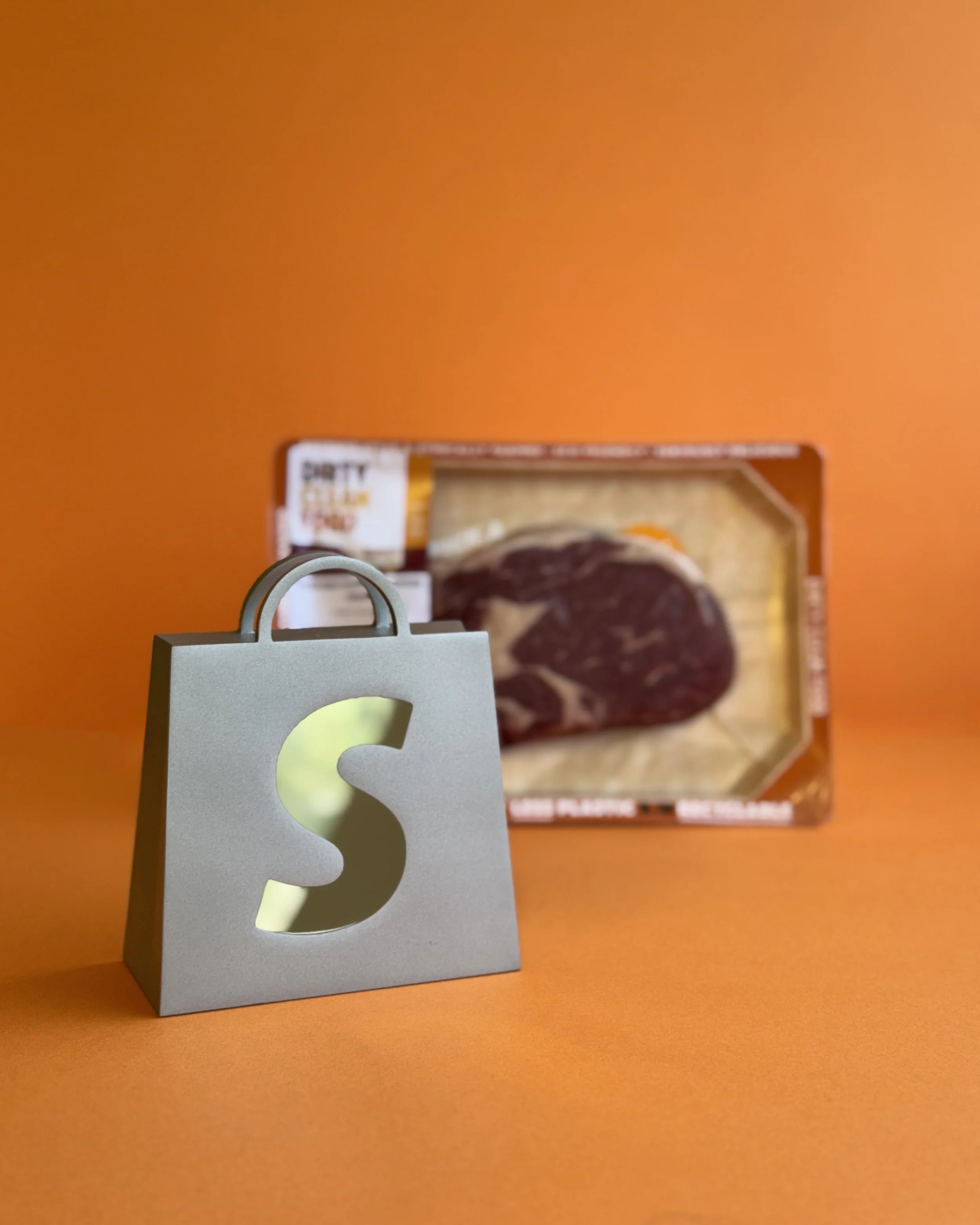 A small gray shopping bag with a cut-out 'S' on the front, in front of a package of raw steak on an orange background.